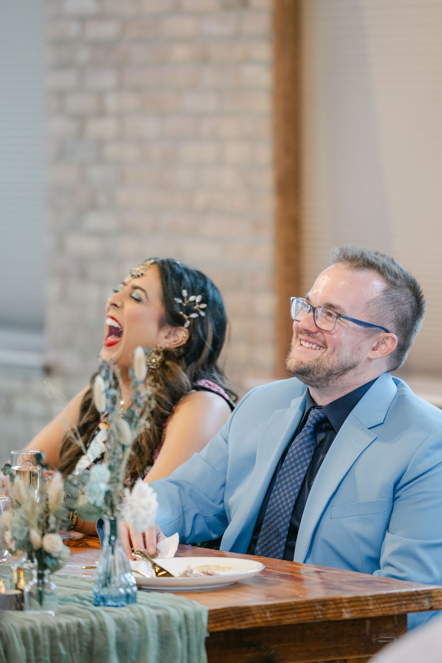 Two people share a joyful moment of laughter while sitting at a wooden table. - The Brix on the Fox
