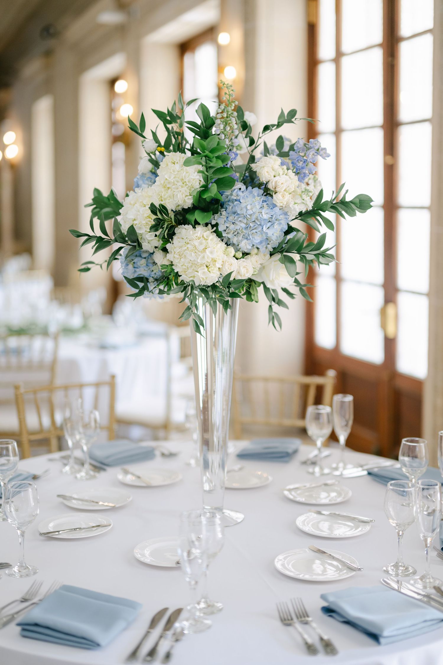 Elegant centerpiece of blue and white hydrangeas in tall glass vase on formal table setting.