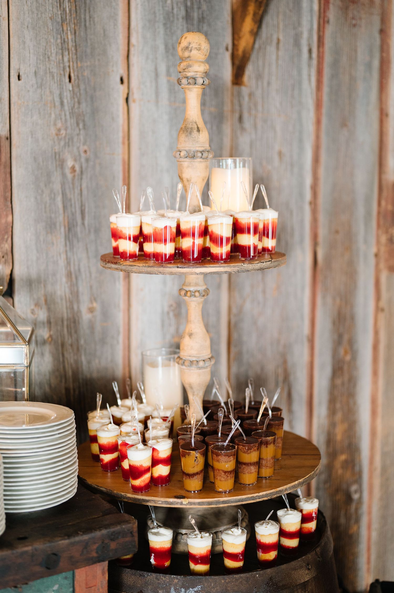 Three-tiered rustic wooden dessert stand displaying colorful layered pudding shots and beverages at a party.