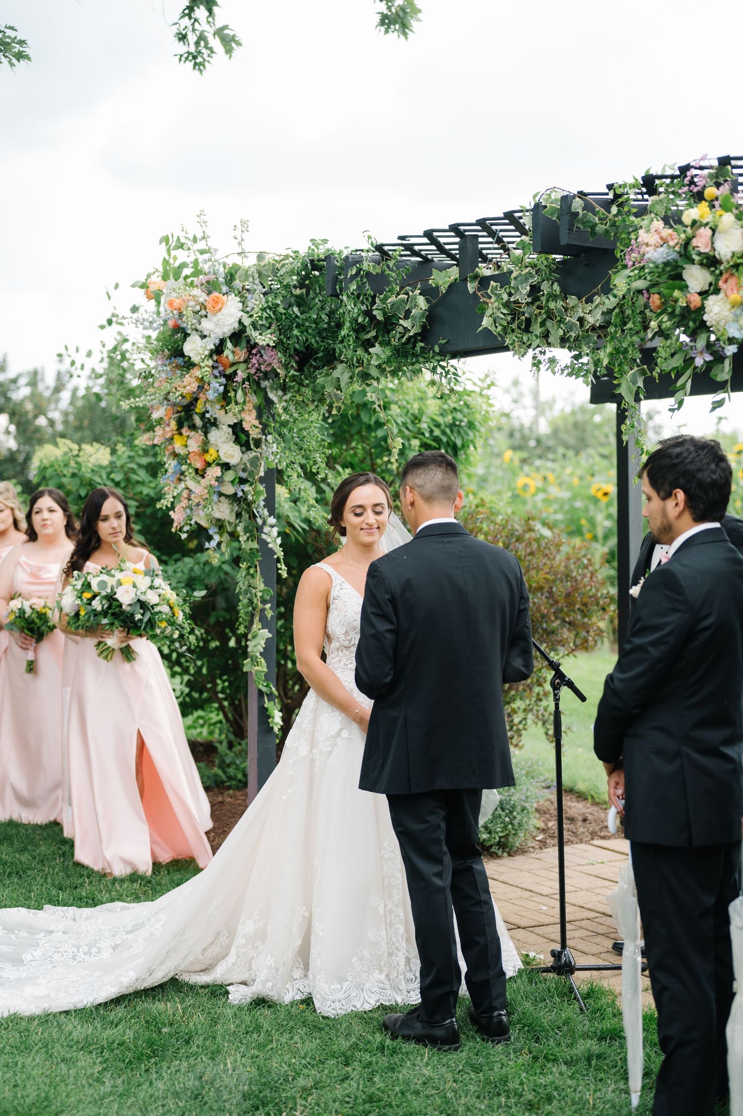 Outdoor wedding ceremony under a floral arch with wedding party in blush and black attire. - Heritage Prairie Farm