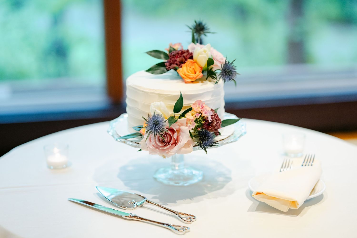 A small two-tiered white wedding cake decorated with fresh pink and peach flowers sits on a glass cake stand. - Hyatt Lodge Oak Brook