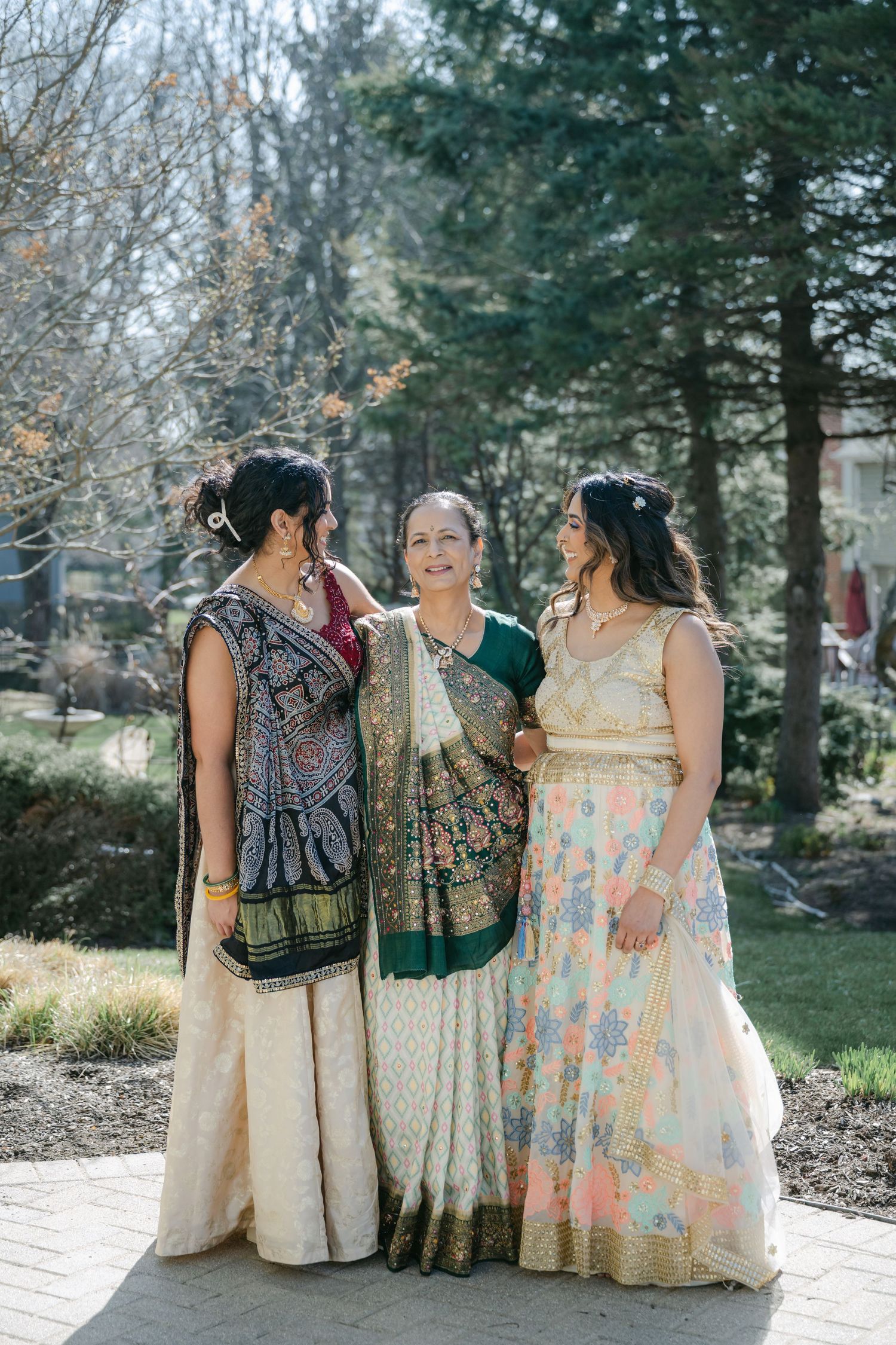 The Brix on the Fox Wedding - Three people in vintage-style dresses sit together on stone steps in a garden setting during a sunny day.