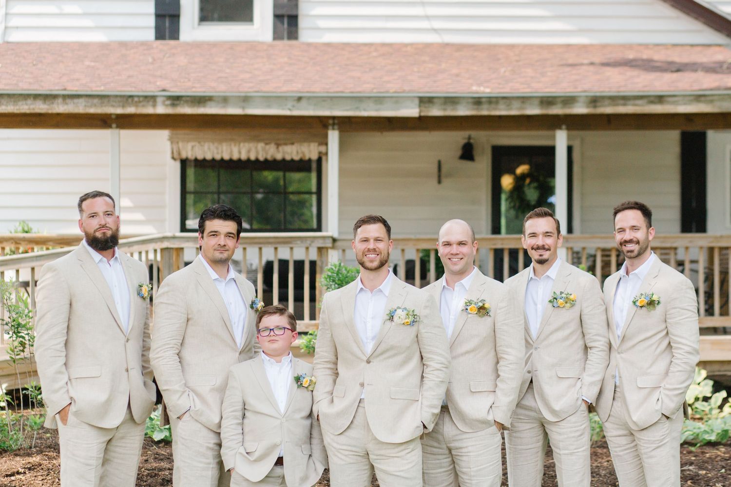 A wedding party in light beige suits stands together on a porch of a white house.