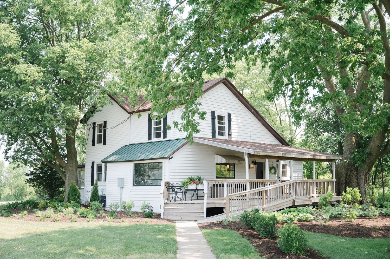 A charming white farmhouse with a wraparound porch nestled among lush green trees and landscaping.