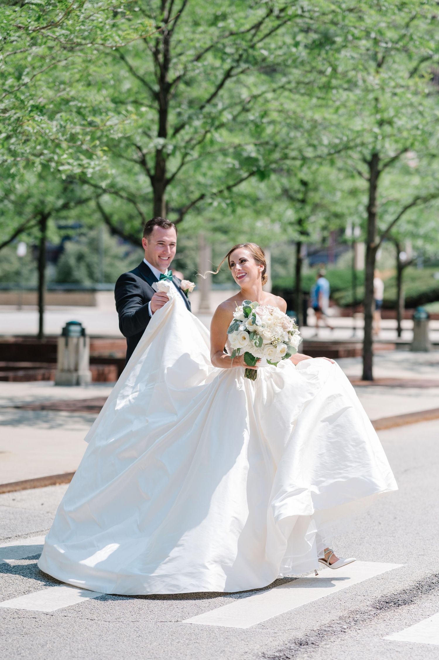 Couple in wedding attire walk together on city street with green trees in background.