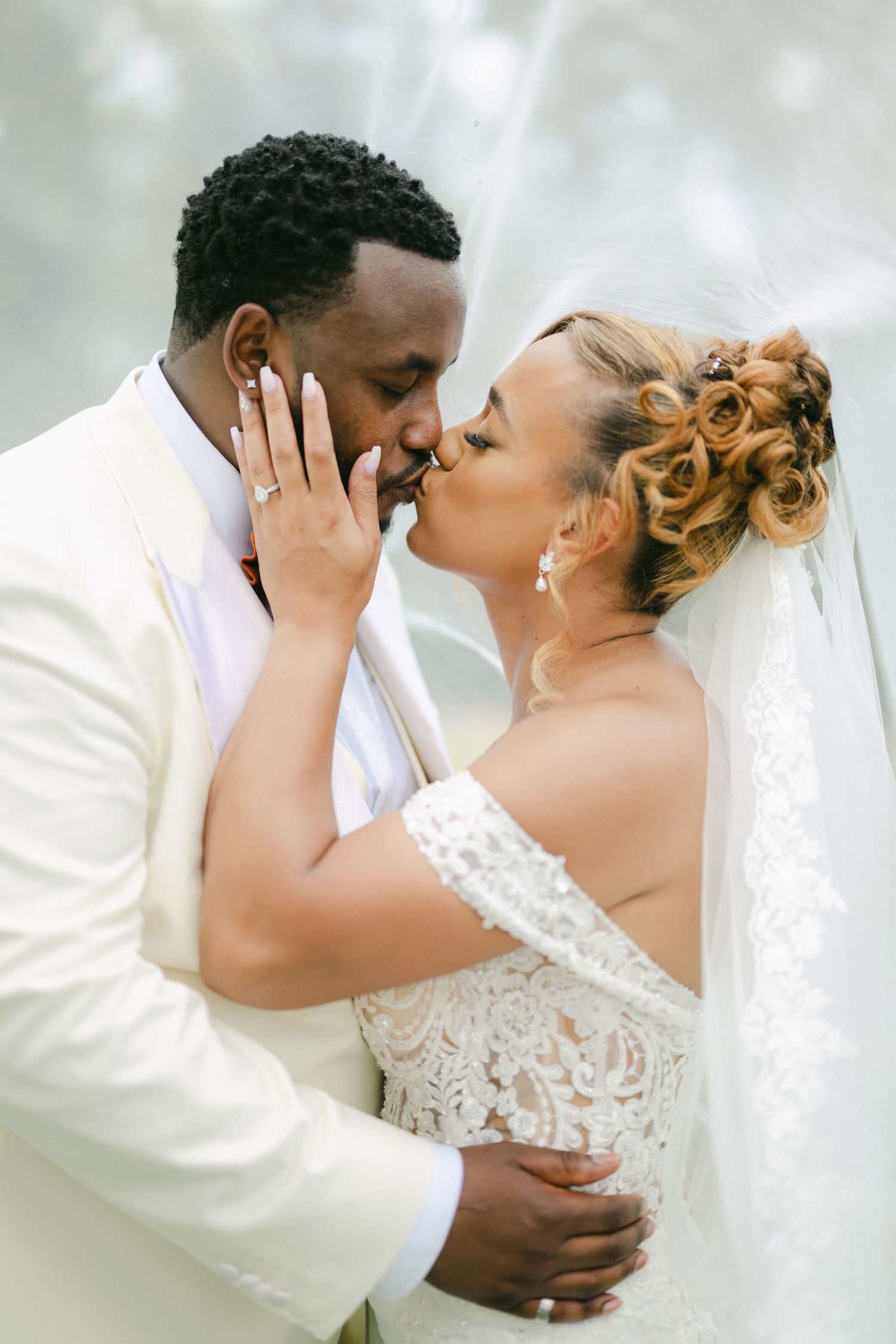 A tender wedding moment with newlyweds sharing an intimate kiss in white formal attire against a dreamy backdrop. - Bird Haven Greenhouse