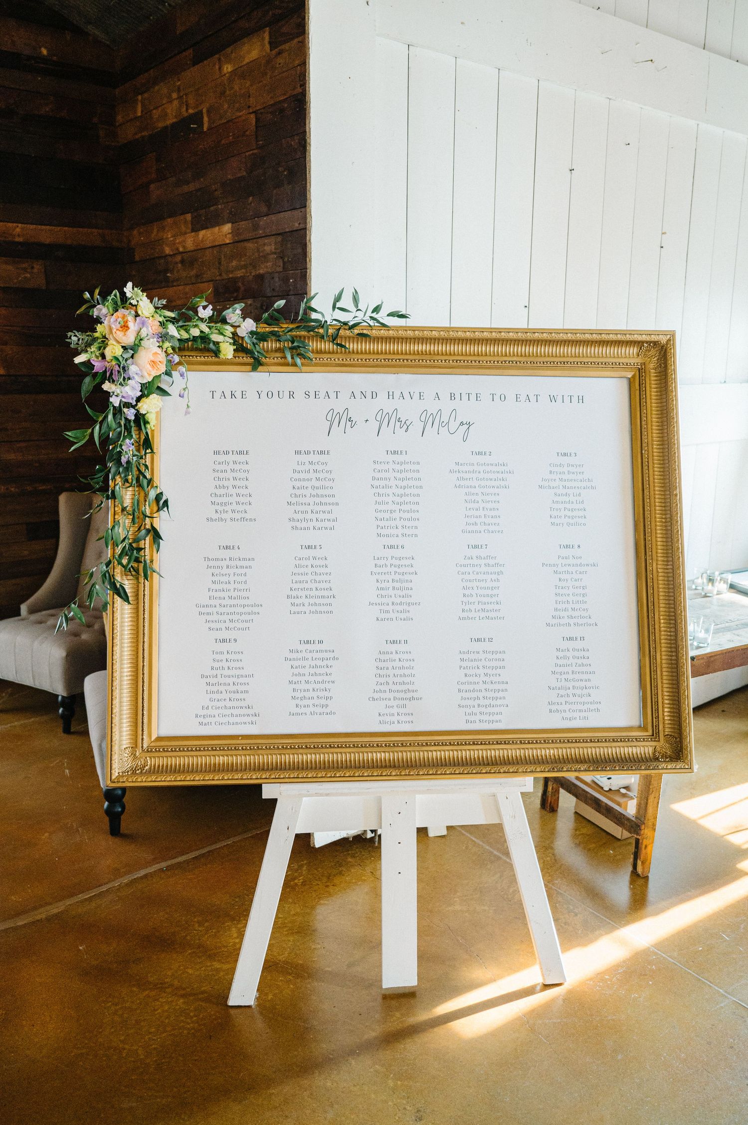 Wedding seating chart displayed in an elegant gold frame decorated with floral accents on a white easel in rustic venue.