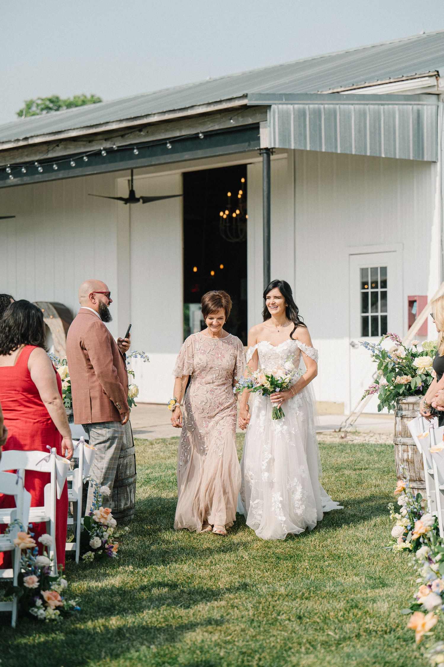 A wedding ceremony processional takes place outside a white farmhouse venue with string lights and floral decorations.