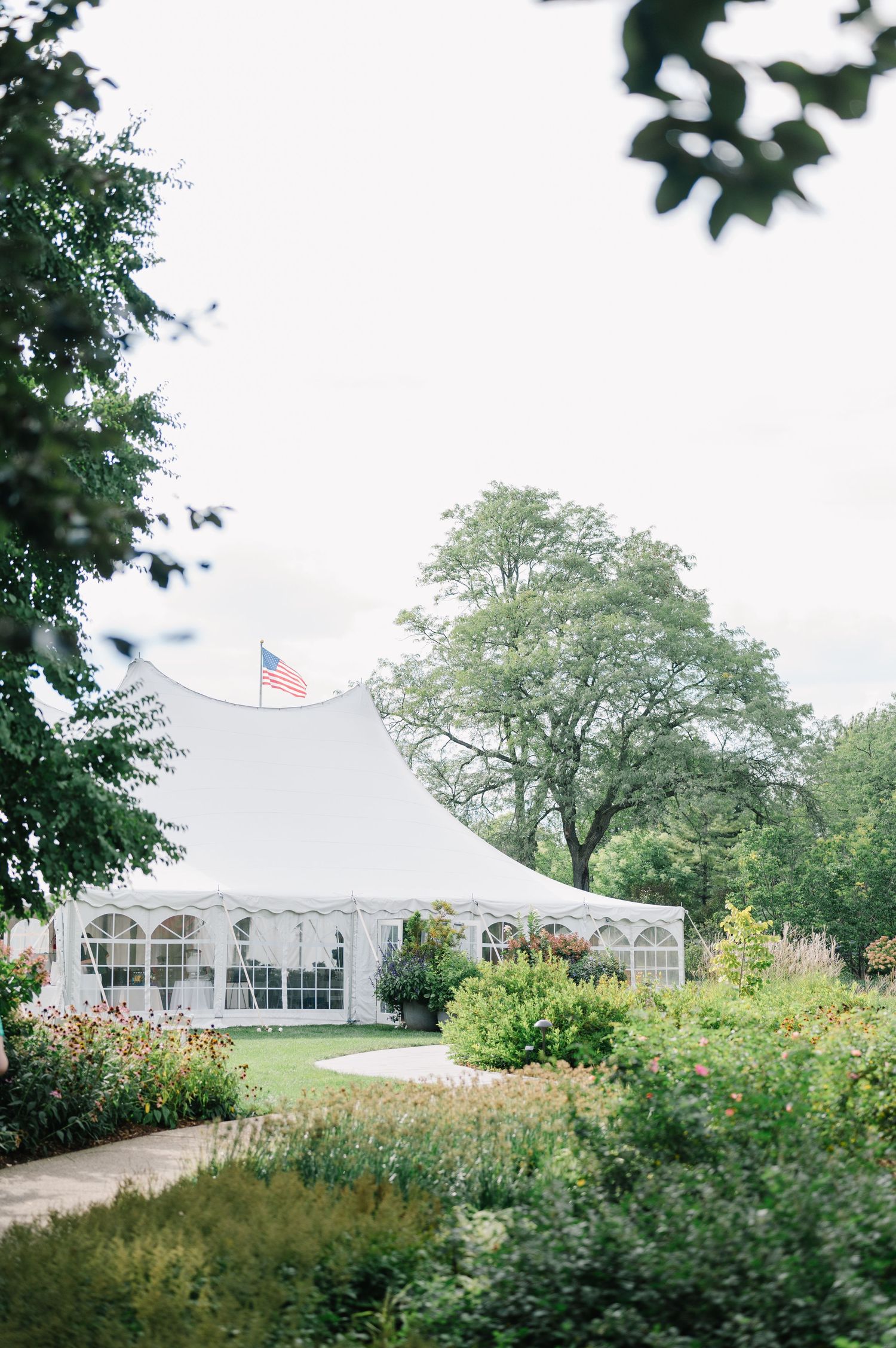 A white wedding tent with an American flag stands among lush gardens and trees on a cloudy day. - Cantigny Park