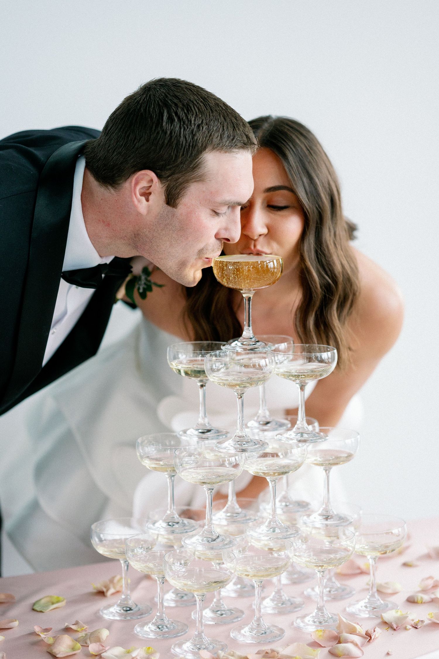 A couple leans in to sip champagne from a tower of delicate coupe glasses adorned with rose petals.