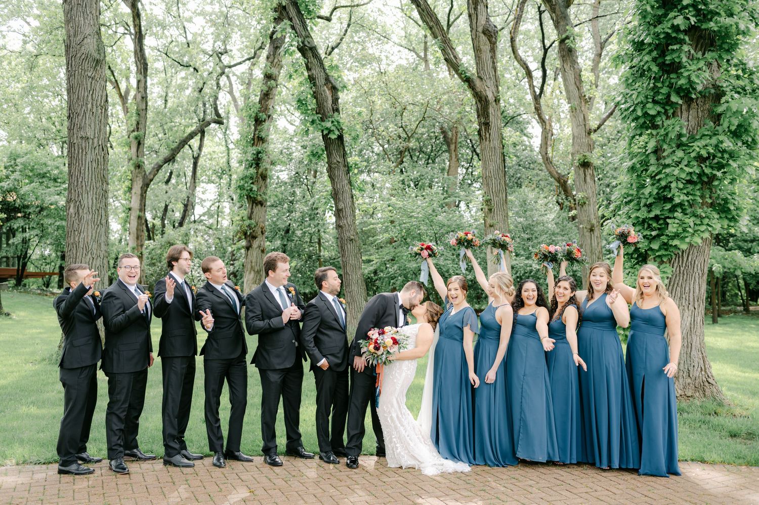 Wedding party in formal attire poses together in a line against a backdrop of lush green trees in a park setting. - Hyatt Lodge Oak Brook