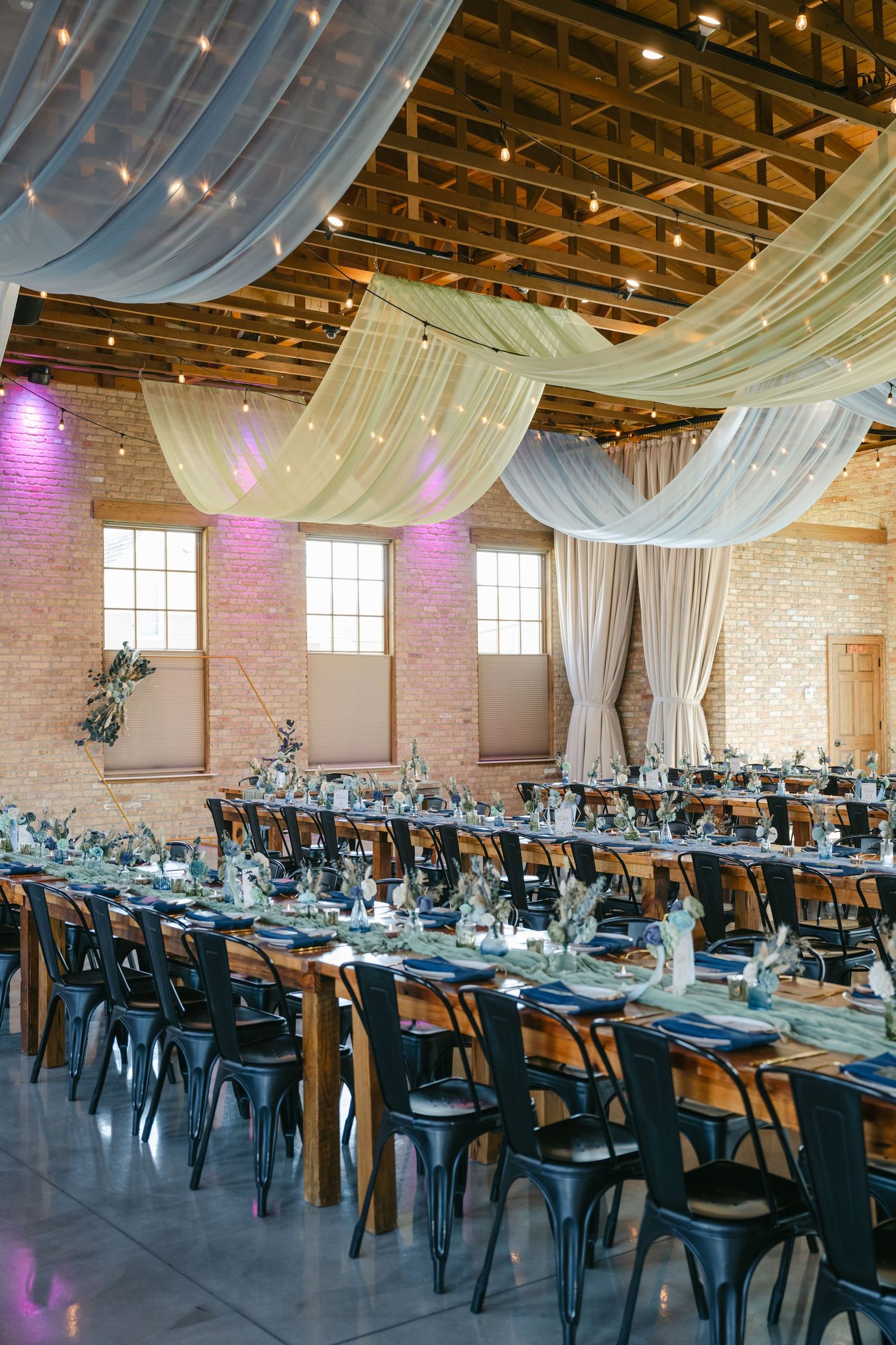 Wedding reception venue with exposed brick walls, white ceiling drapes, and blue table settings. - The Brix on the Fox