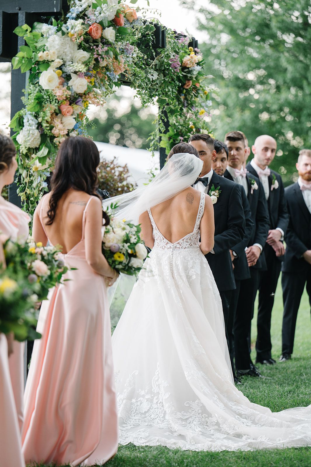 Bride in lace backless gown with veil at outdoor wedding ceremony beneath floral arch. - Heritage Prairie Farm Wedding