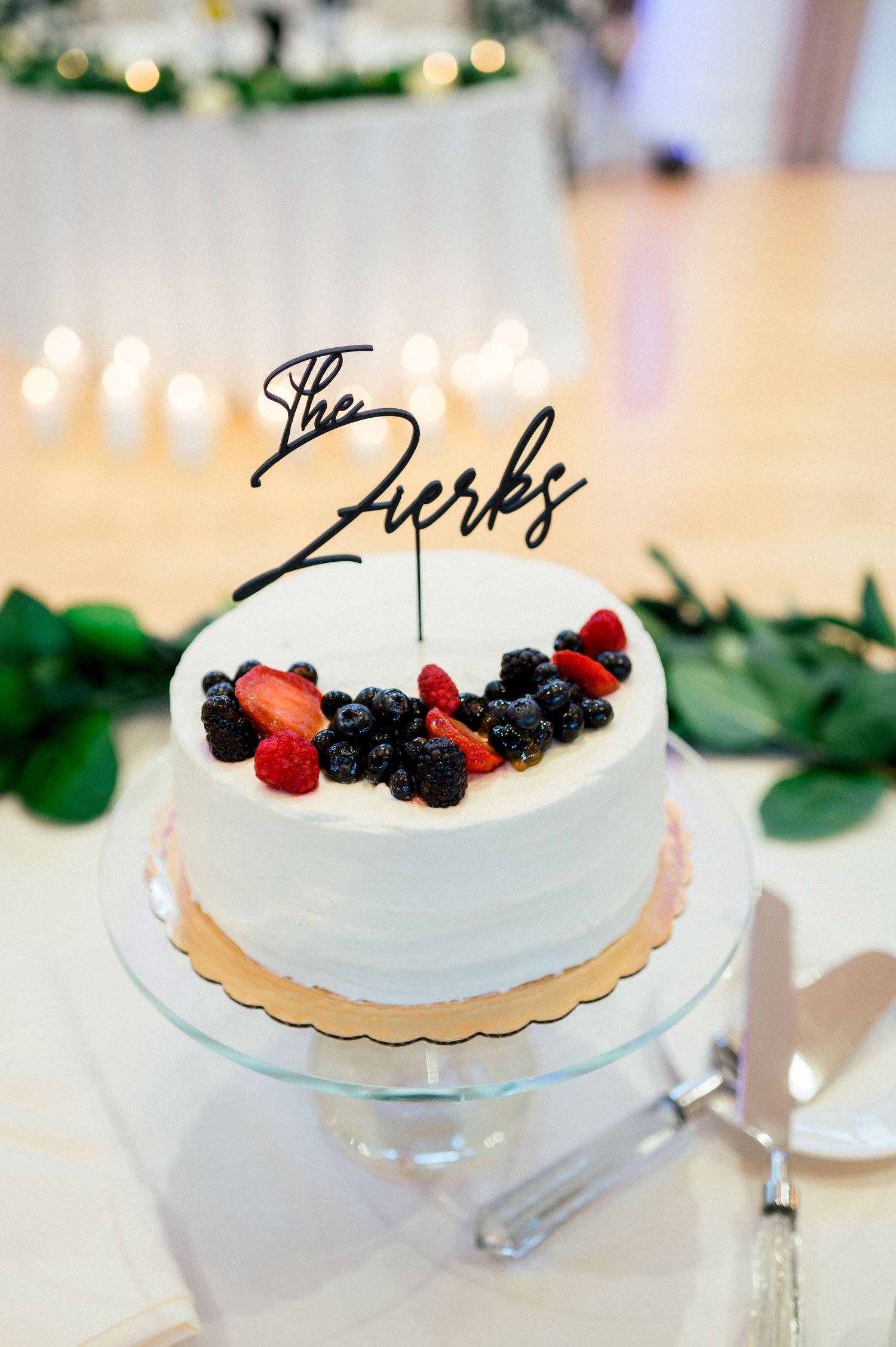 White frosted cake decorated with fresh berries and greenery on elegant cake stand at wedding reception.