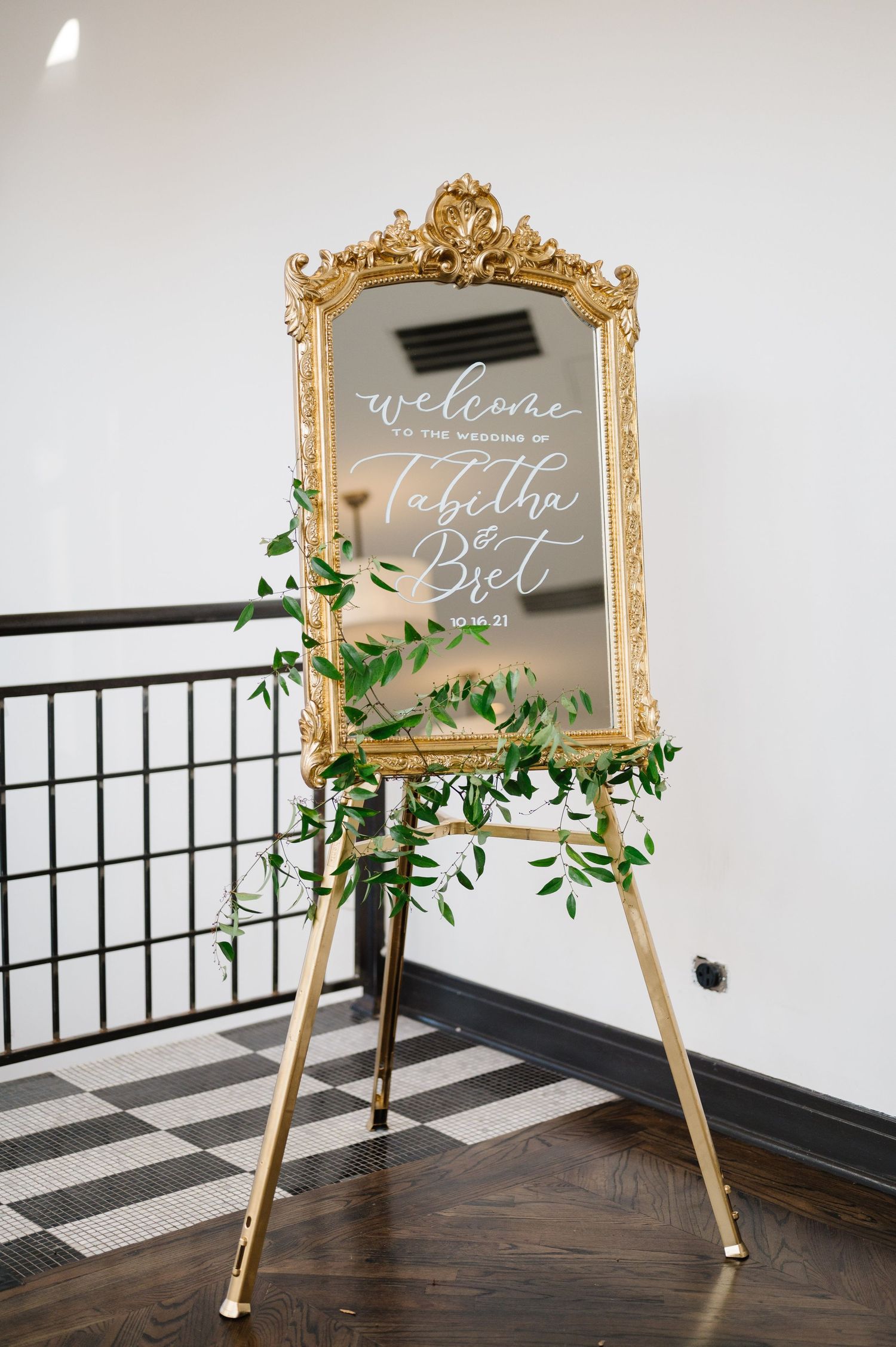 Elegant gold ornate mirror with wedding welcome text stands on wooden easel decorated with greenery vines. - Mae District Chicago