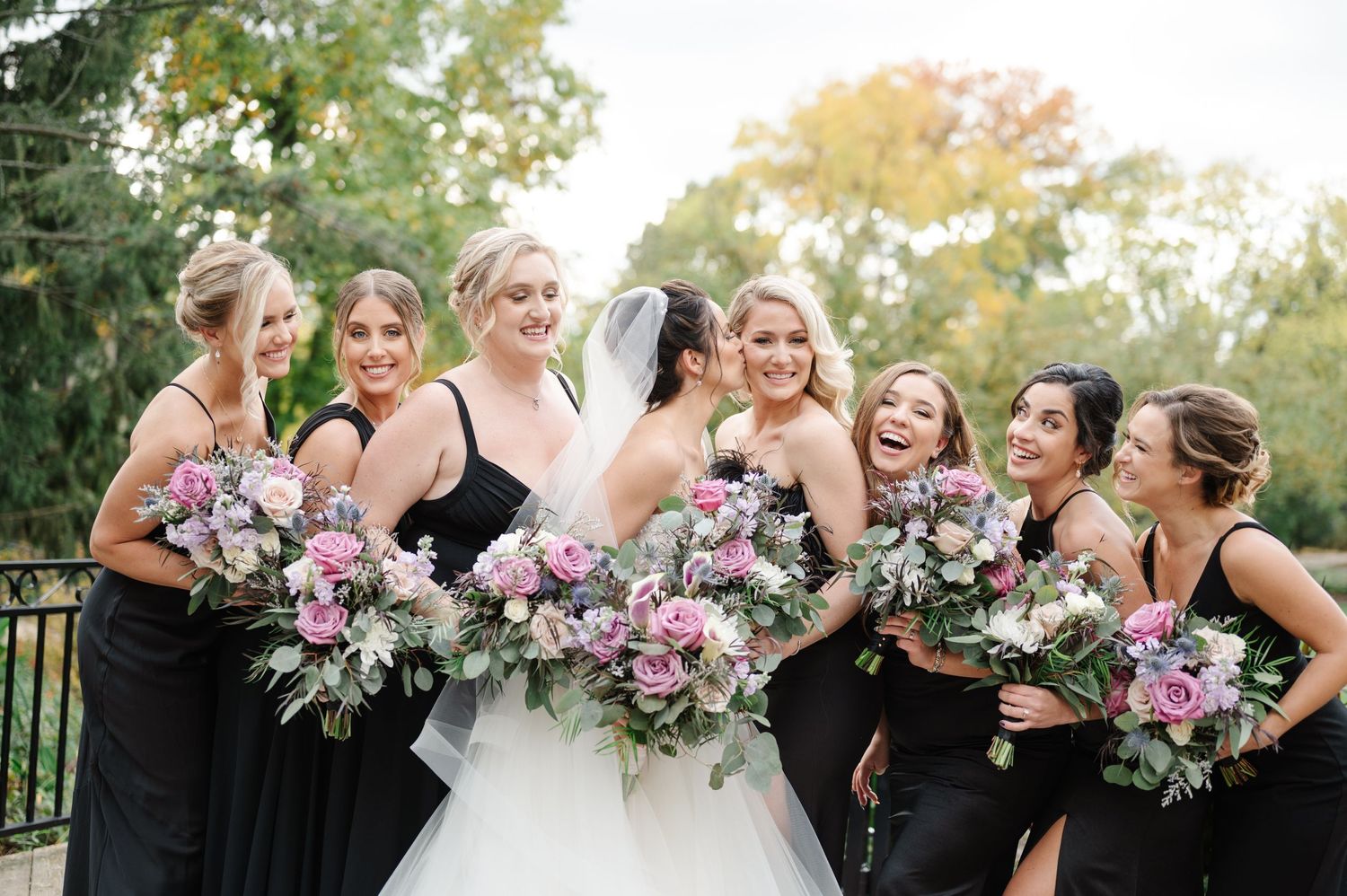 Wedding party in black bridesmaid dresses share a candid laughing moment while holding pink and purple floral bouquets.