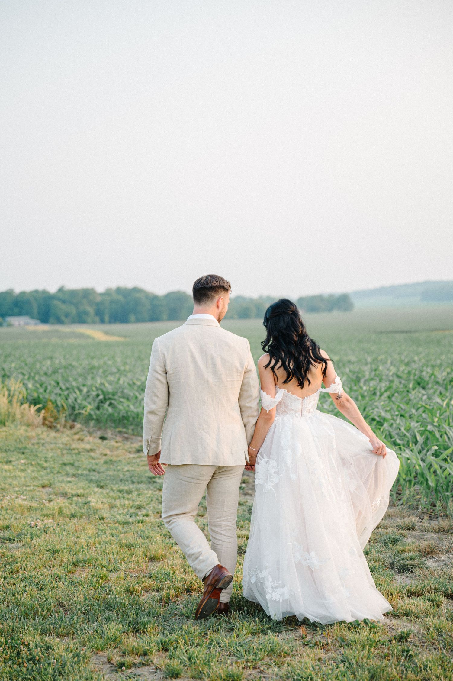 Wedding couple walks through field, bride wearing white lace dress and groom in beige suit. - Venue 5126 Wedding