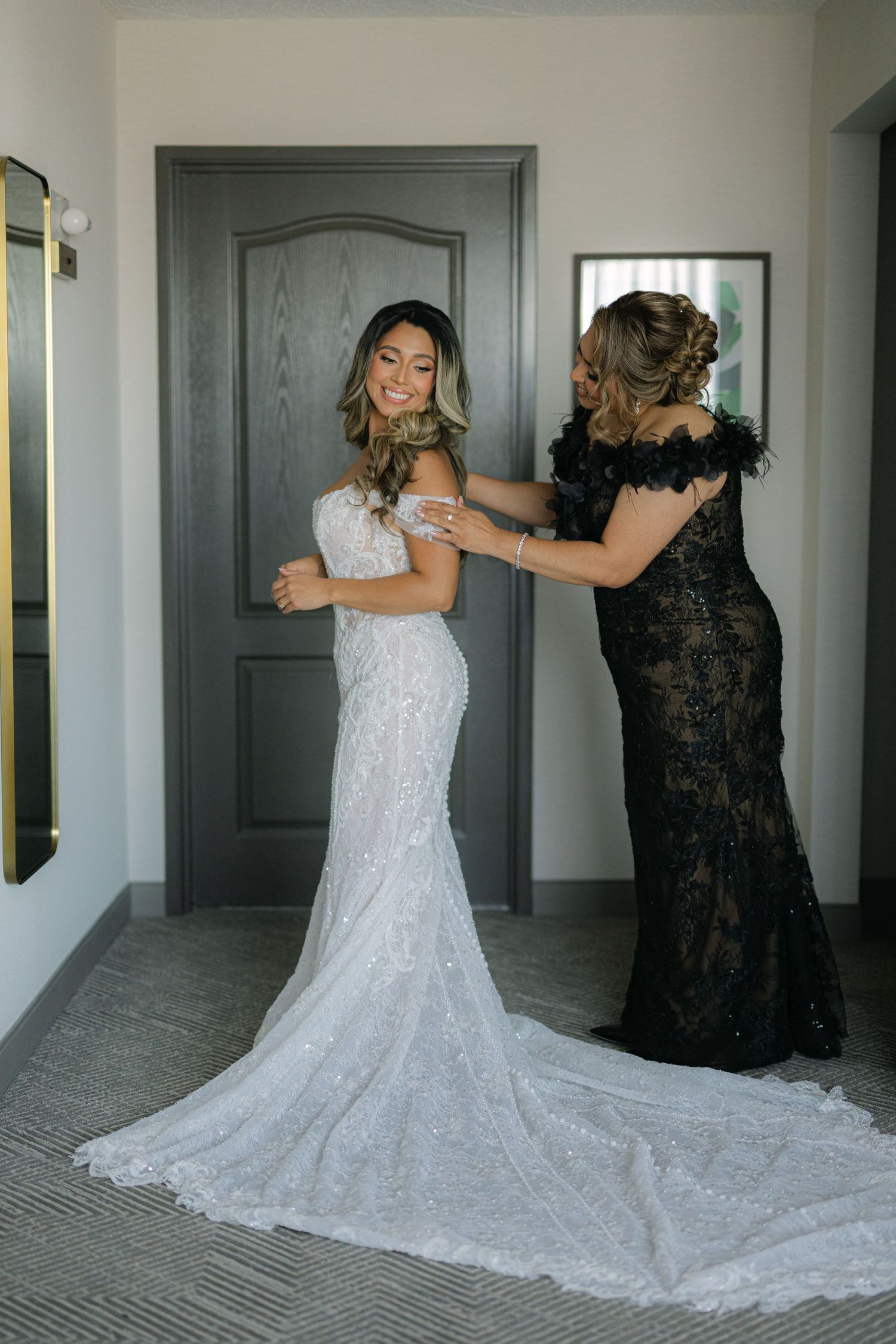 A bride in a white lace wedding gown with long train gets help with her dress from someone wearing a black formal gown.