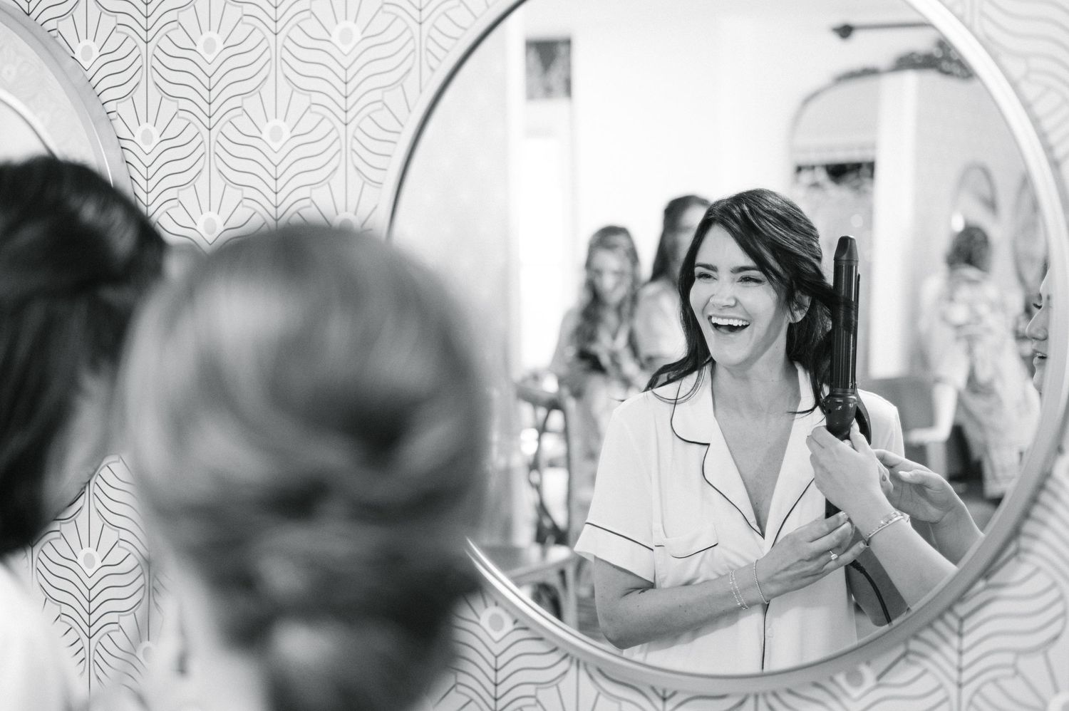 A black and white reflection in a round mirror shows a joyful woman in a white blazer laughing during a special occasion.