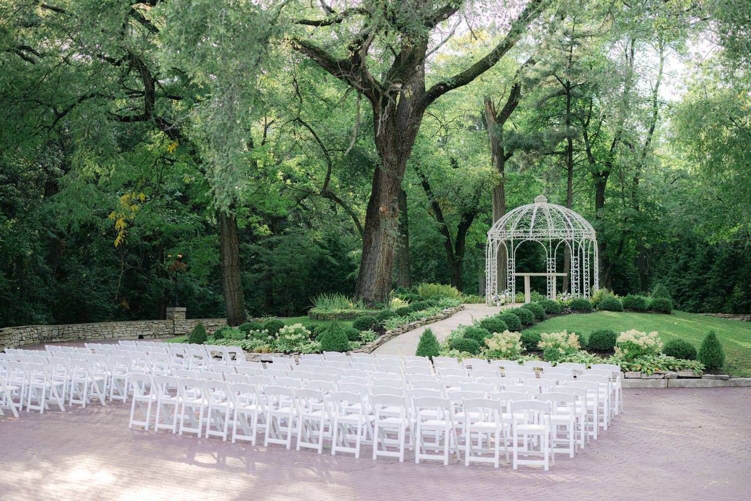 White folding chairs arranged in rows facing an elegant garden gazebo surrounded by lush green trees. - Meyer's Castle Events