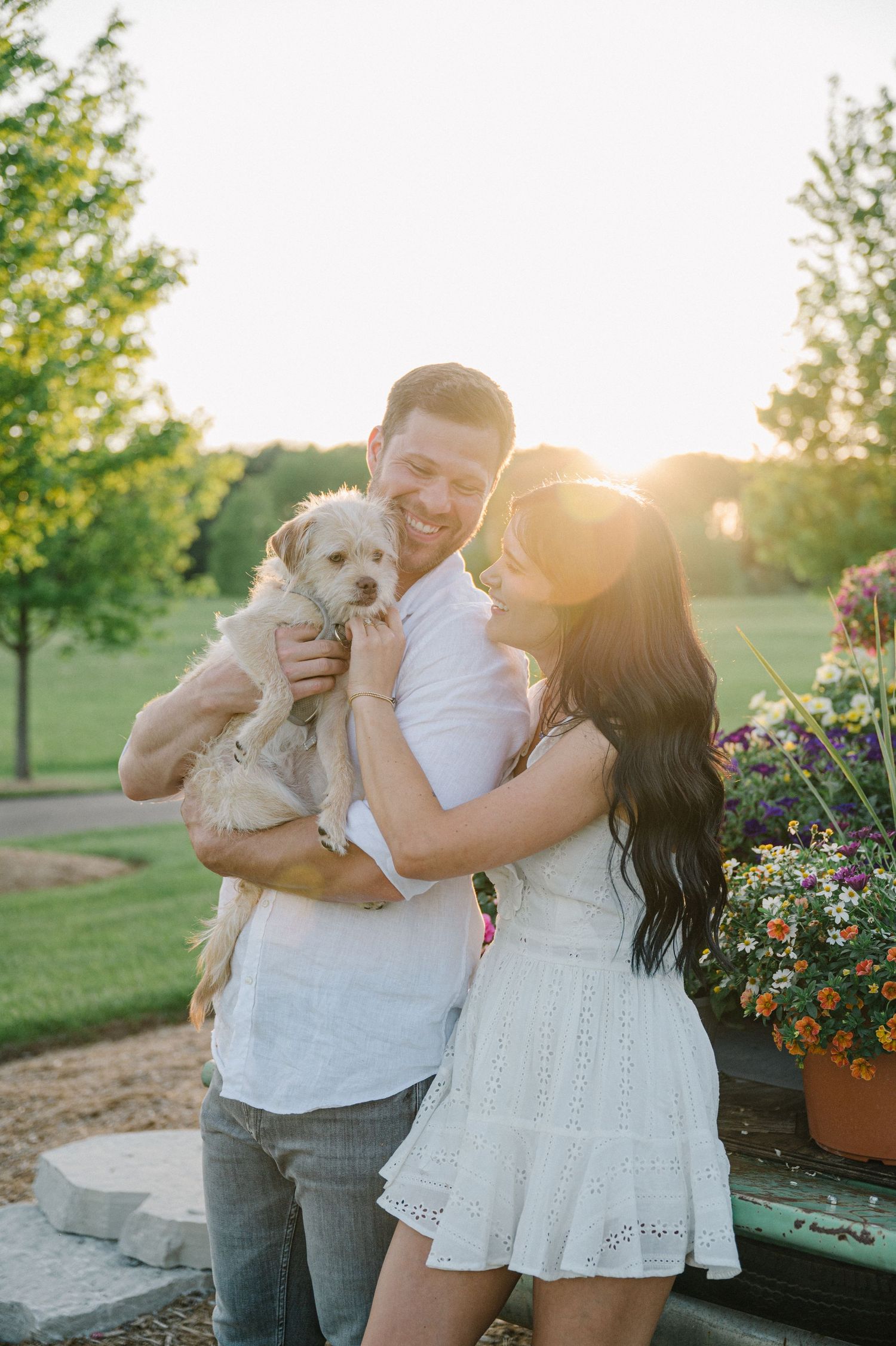 A couple shares tender moments with their small white dog in a garden setting during golden hour sunset. - Oswego, IL