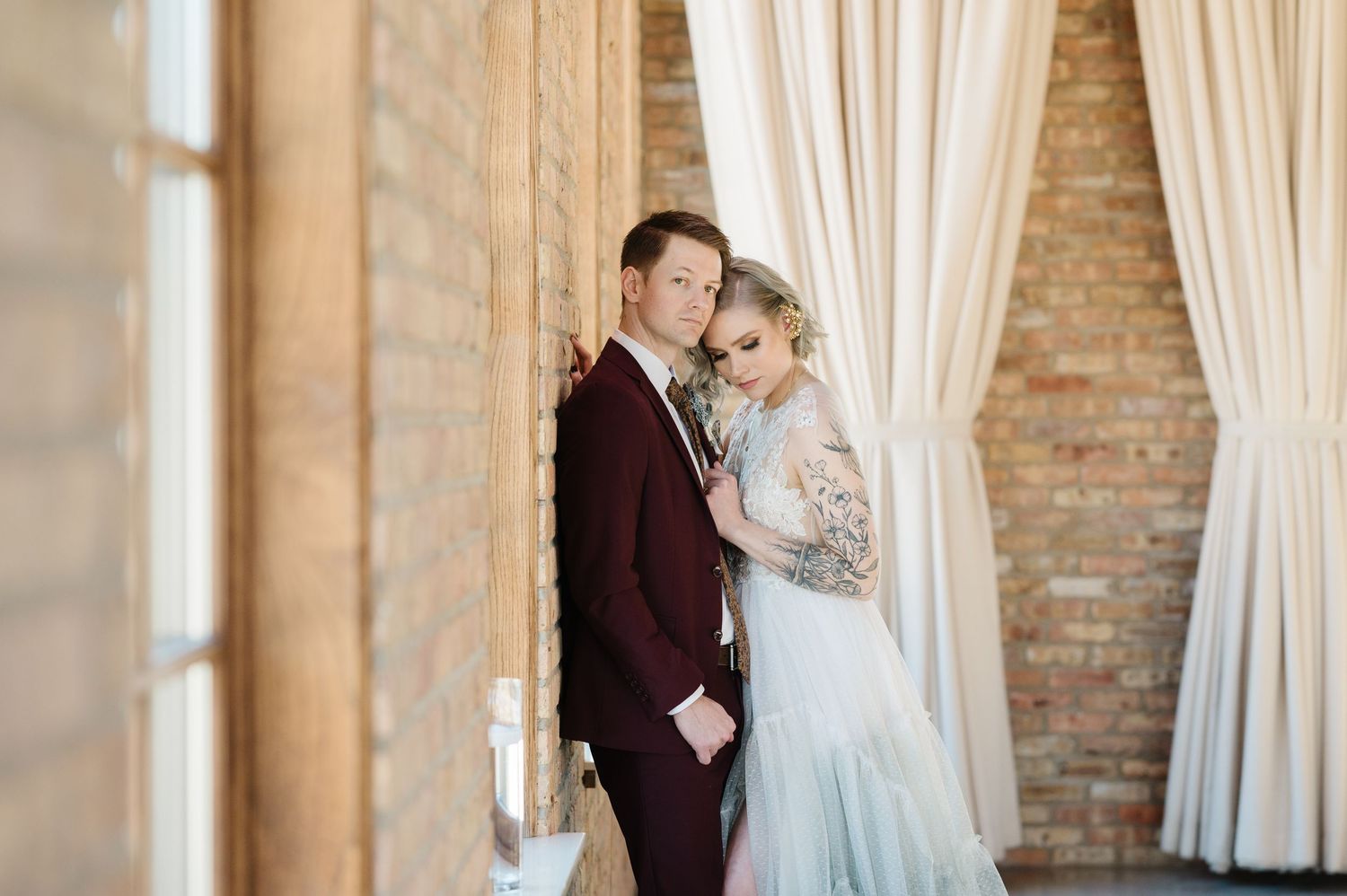 A couple in formal wedding attire stands together near white curtains and exposed brick wall in natural light. - The Brix on the Fox
