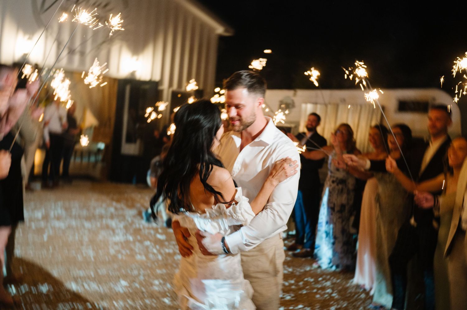 A couple shares their romantic first dance surrounded by wedding guests holding sparklers at an indoor reception.
