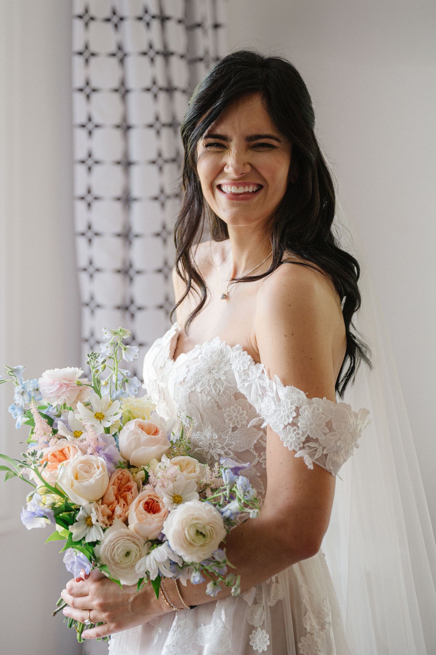 A radiant bride in a lace off-shoulder wedding gown holds a romantic bouquet of blush and white roses and delphinium.