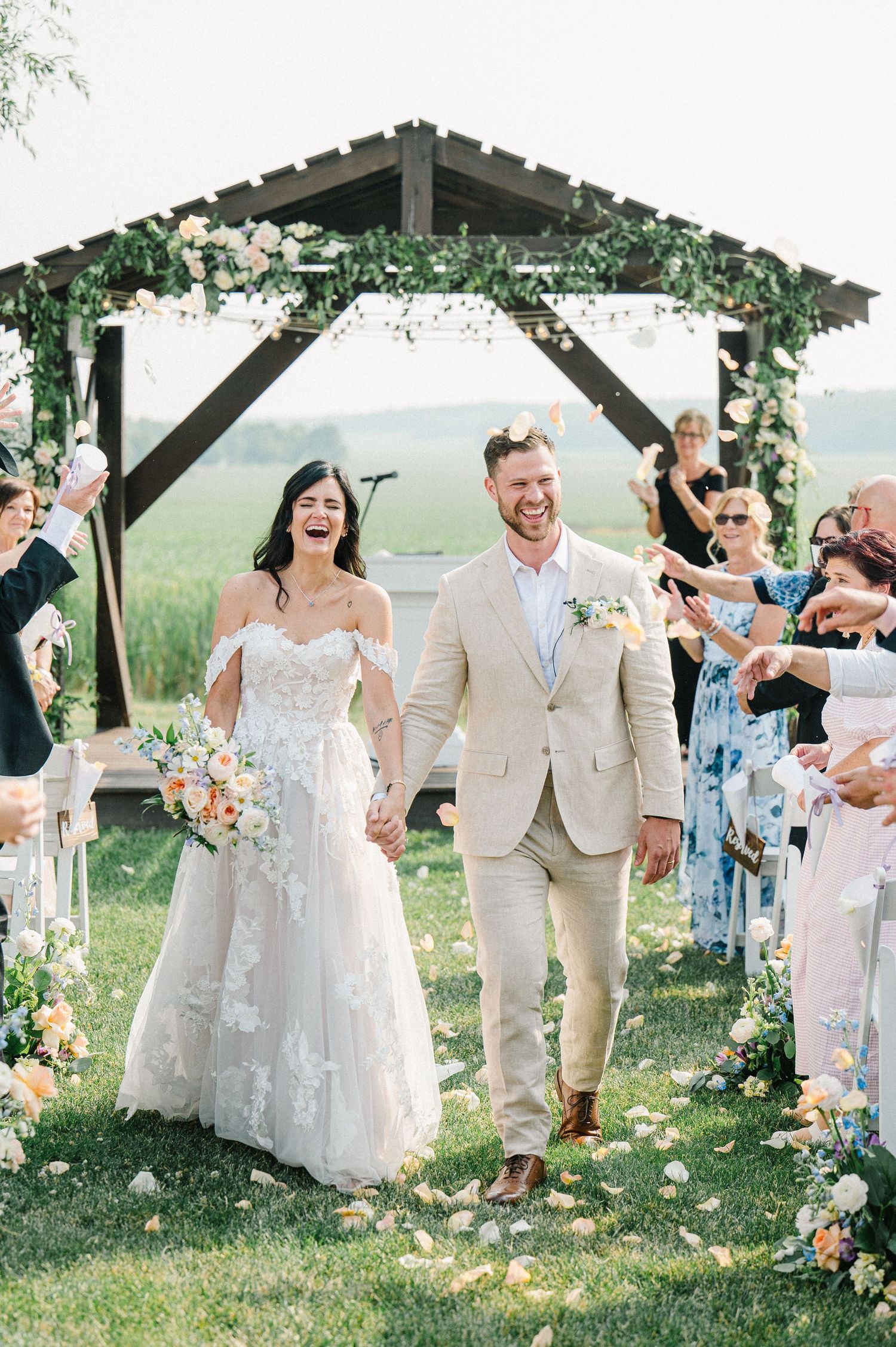A joyful outdoor wedding ceremony under a wooden pergola decorated with florals and greenery in a scenic countryside setting.