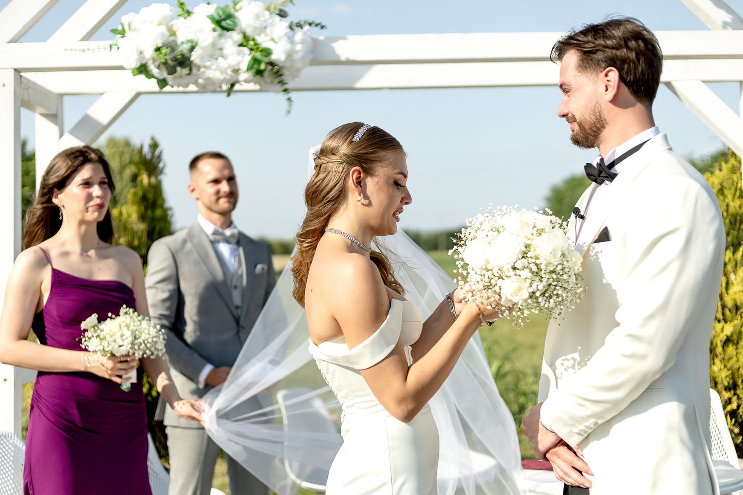 Wedding party members interact during outdoor ceremony under white canopy.