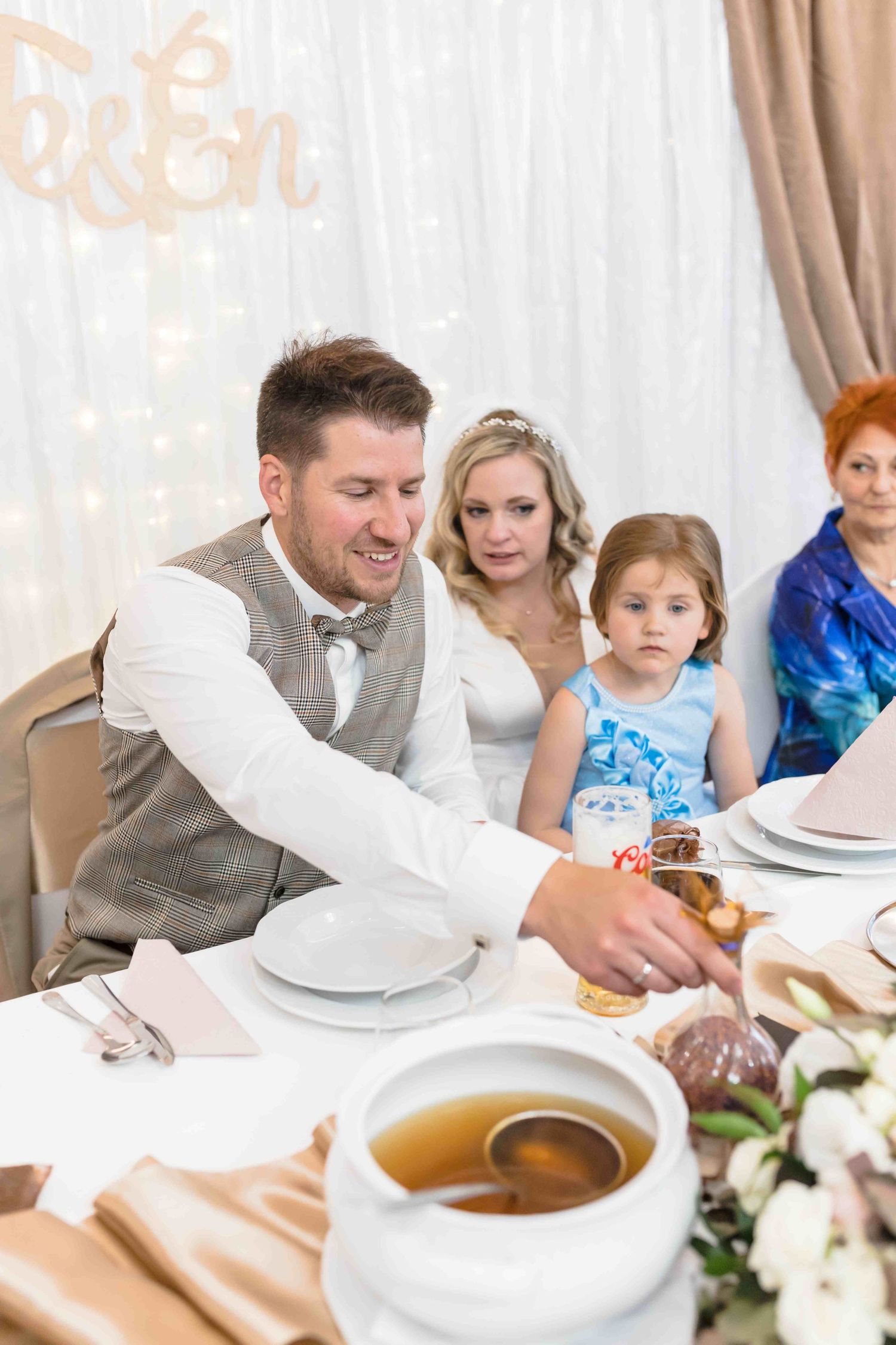 Guests seated at an elegant wedding reception table with a tureen of soup and formal place settings.