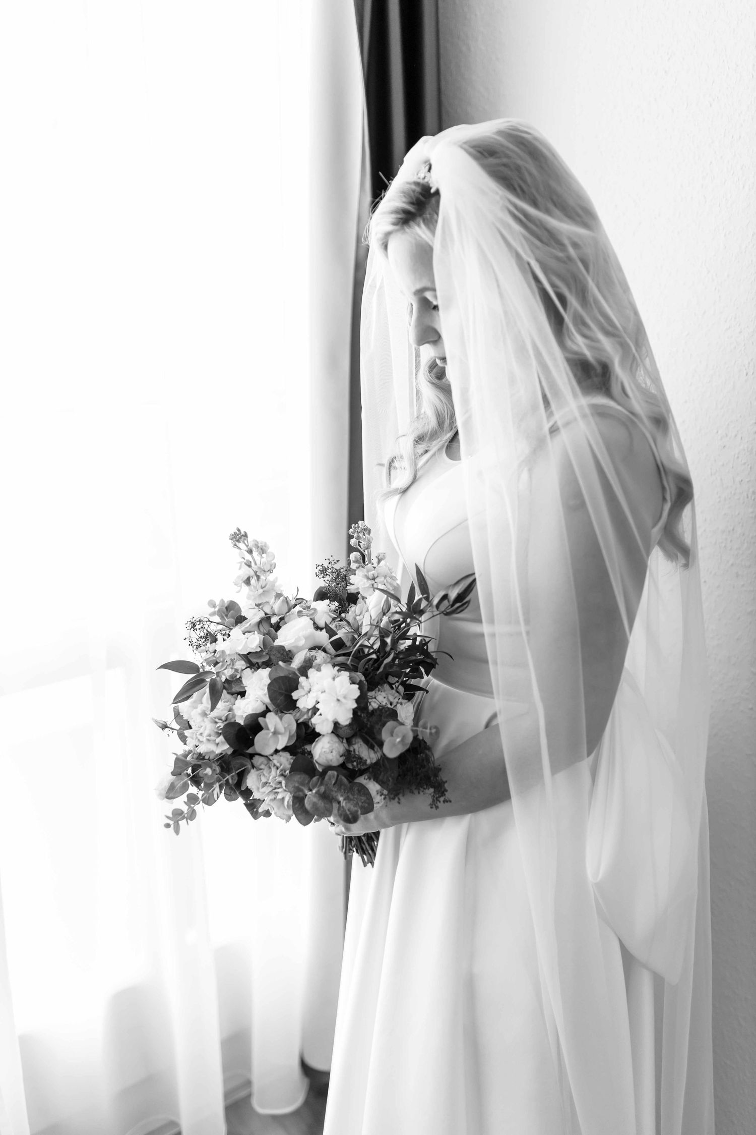 A bride in a white wedding dress and veil stands by a window holding an elegant floral bouquet in black and white.
