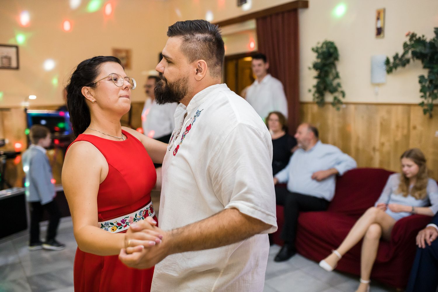 A couple dances together at an indoor venue, with the woman wearing a red sleeveless dress and man in white shirt.