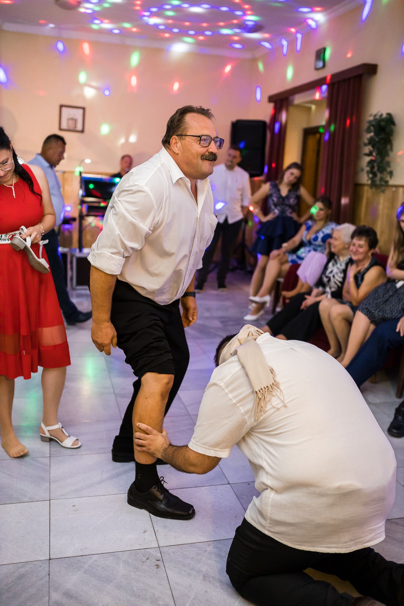 Dancers perform the Macarena dance in a party room with colorful disco lights overhead.