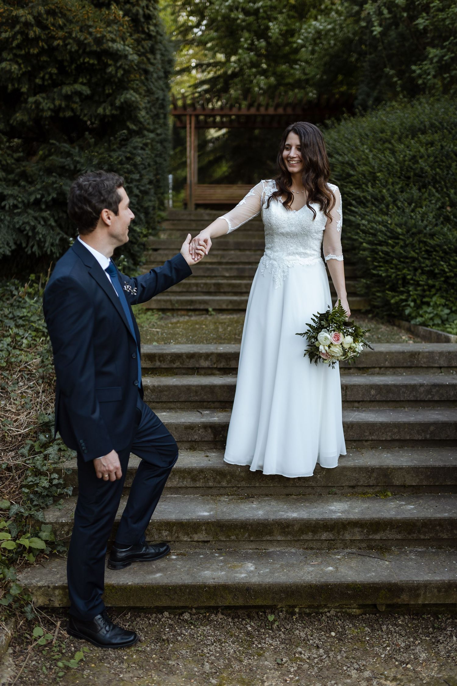 A couple in wedding attire shares a playful moment on outdoor stone steps surrounded by greenery.