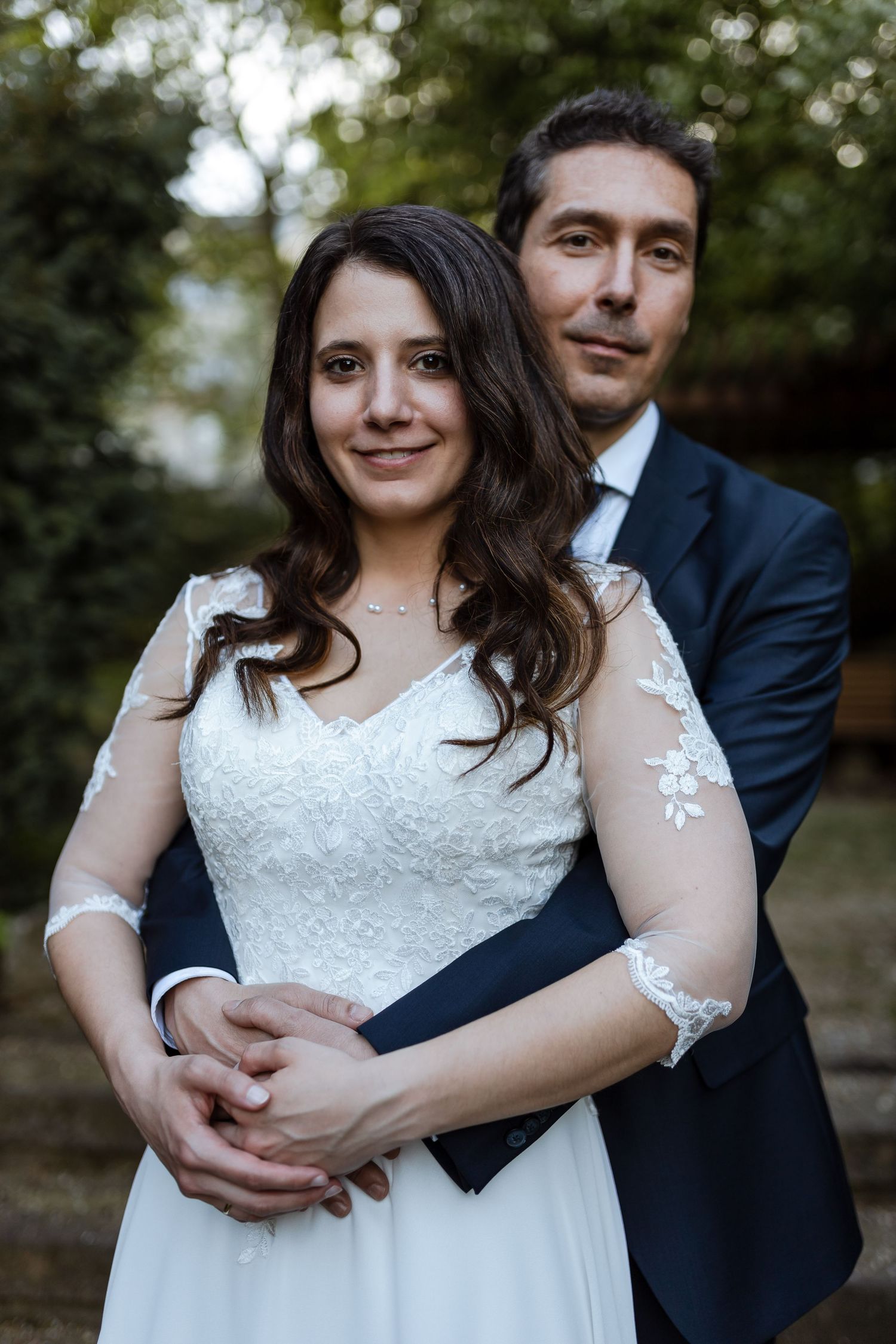 A romantic wedding couple embraces outdoors, with the bride wearing a white lace dress and groom in a navy suit.