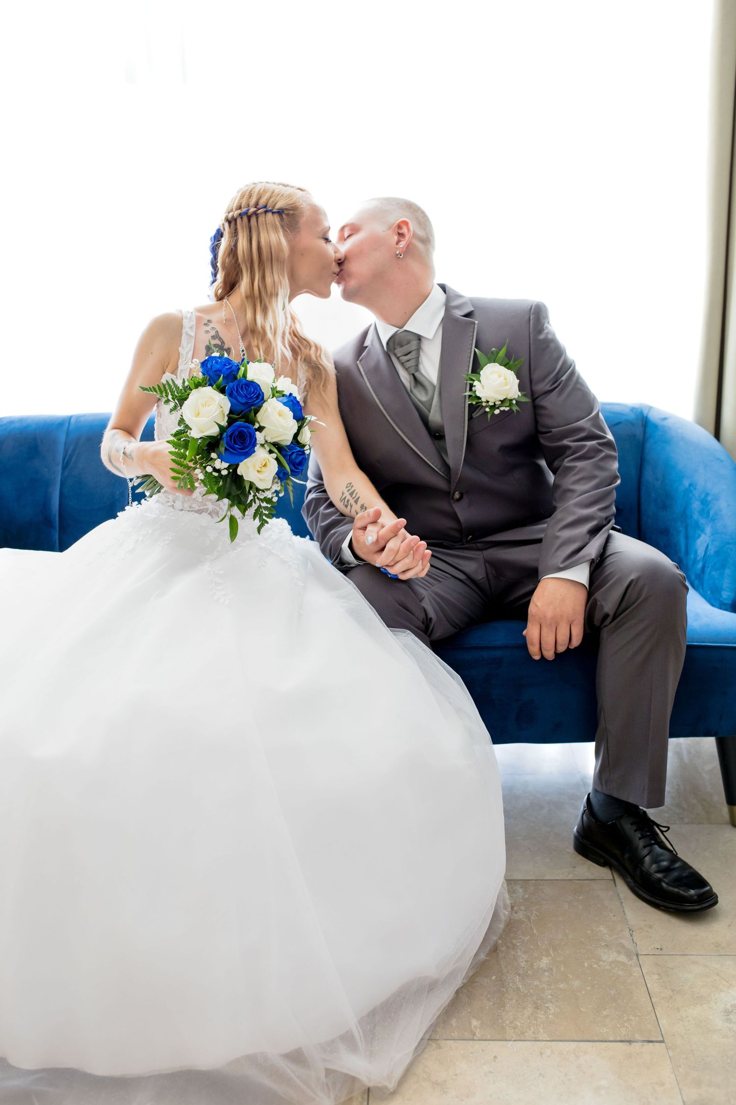 Newlywed couple sharing a kiss on a blue velvet couch, with bride holding a blue and white floral bouquet.