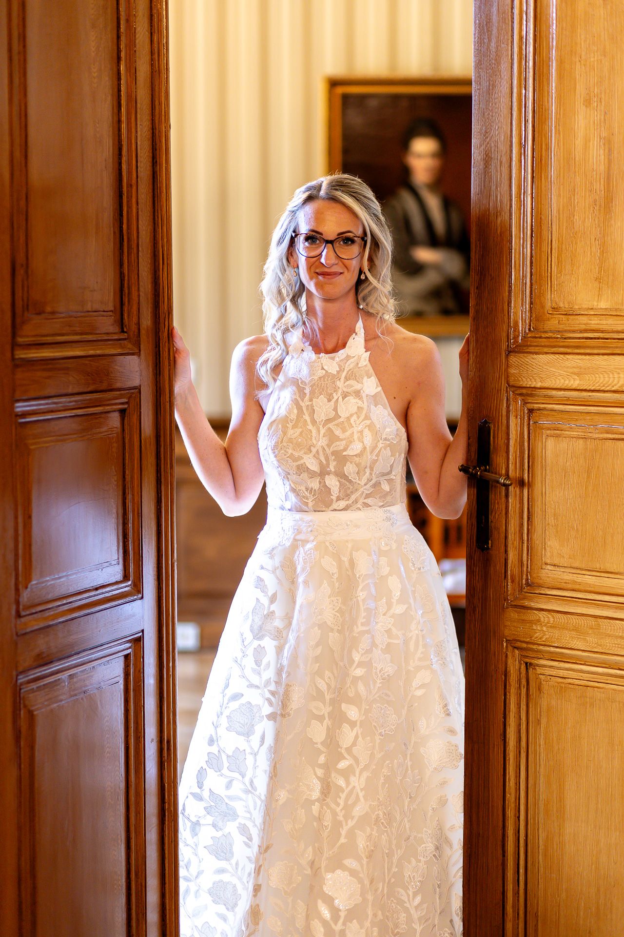 A bride in a white lace halter dress and glasses stands between wooden doors in an elegant interior setting.