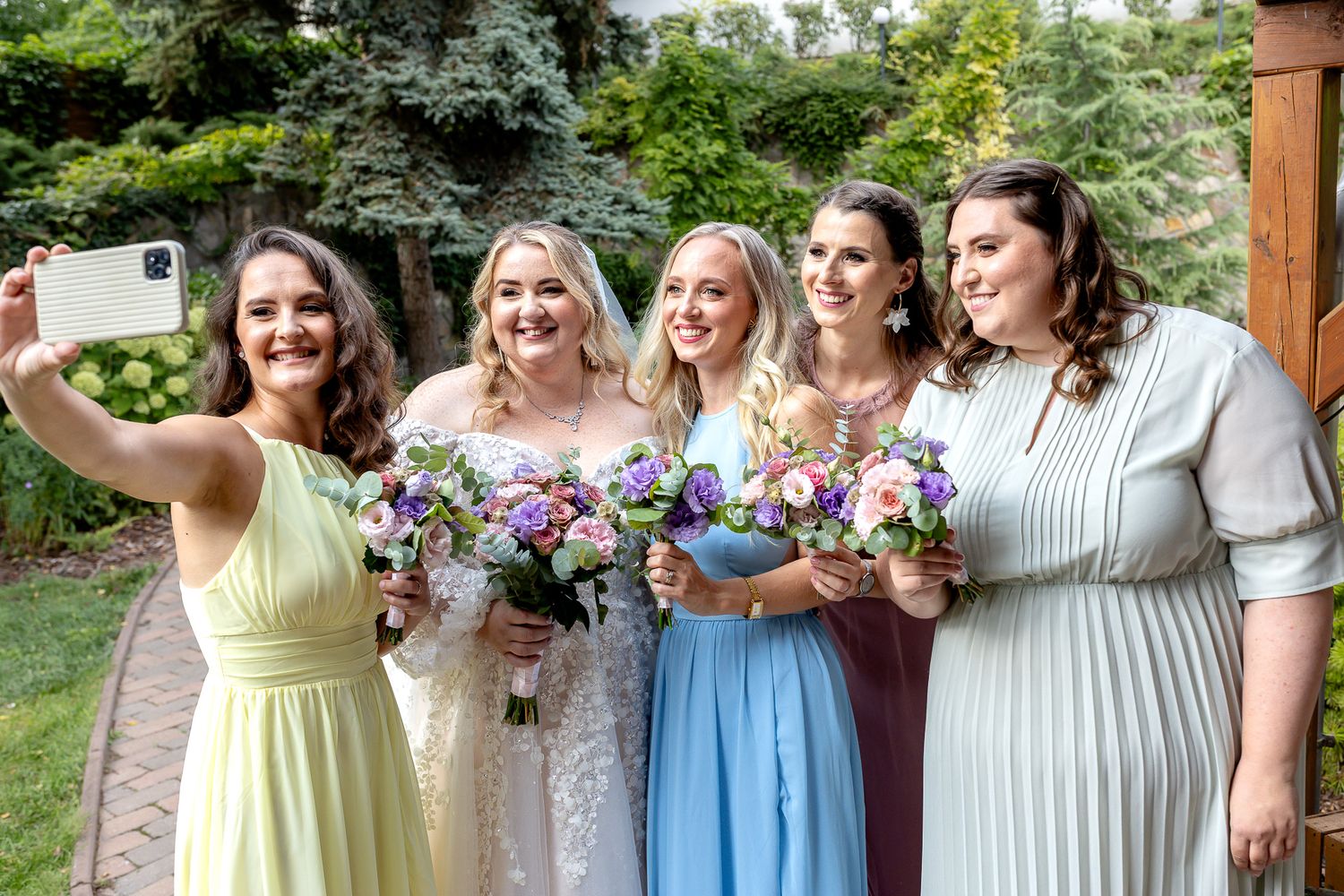 A group in coordinated pastel dresses stands together holding colorful floral bouquets at an outdoor event.