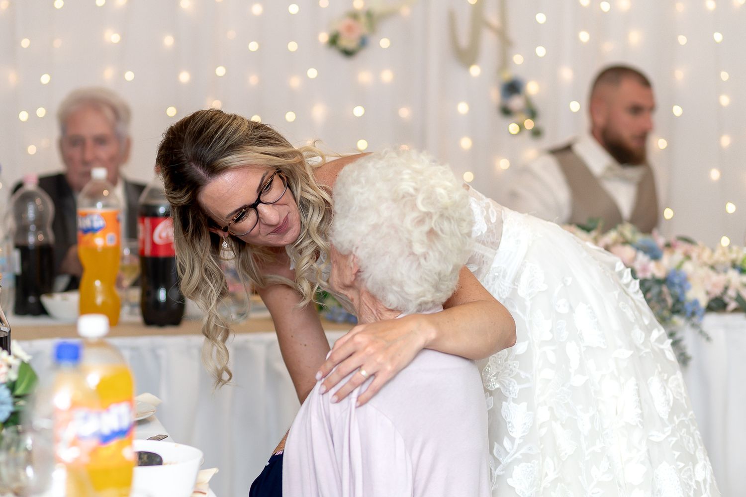 Newlyweds embrace and kiss during their wedding reception celebration with string lights and flowers decorating the venue.