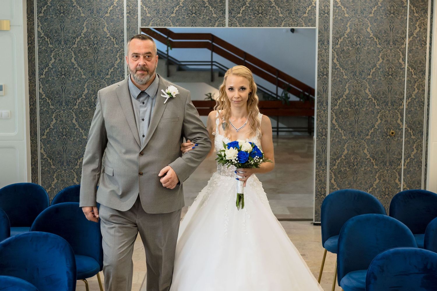 A wedding party walks down a blue carpeted aisle in a modern venue with gray walls and matching attire.
