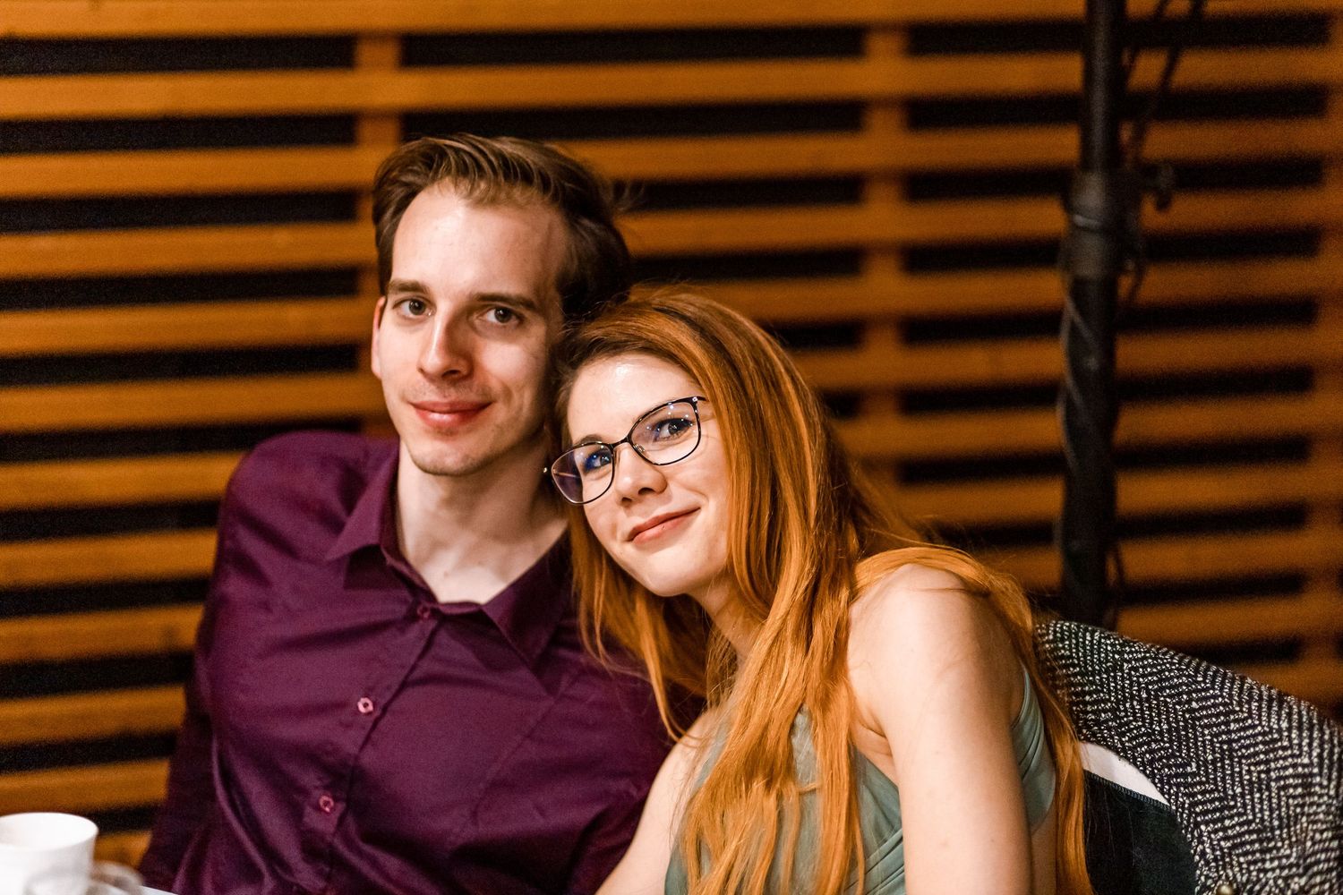 A couple sits close together against a wooden slatted wall, wearing casual attire in warm lighting.