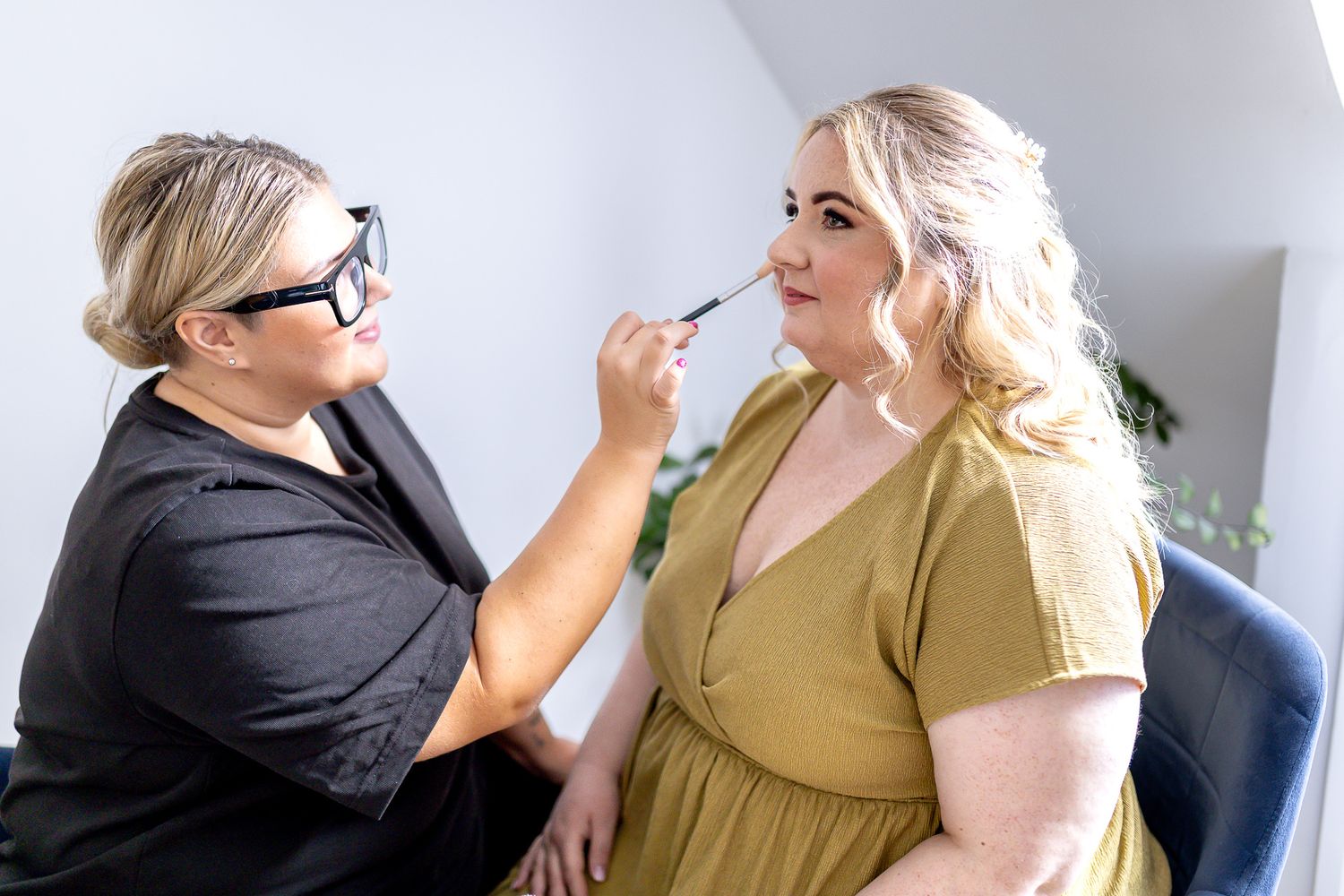 Makeup artist in black applies cosmetics to client wearing olive dress during professional beauty session.