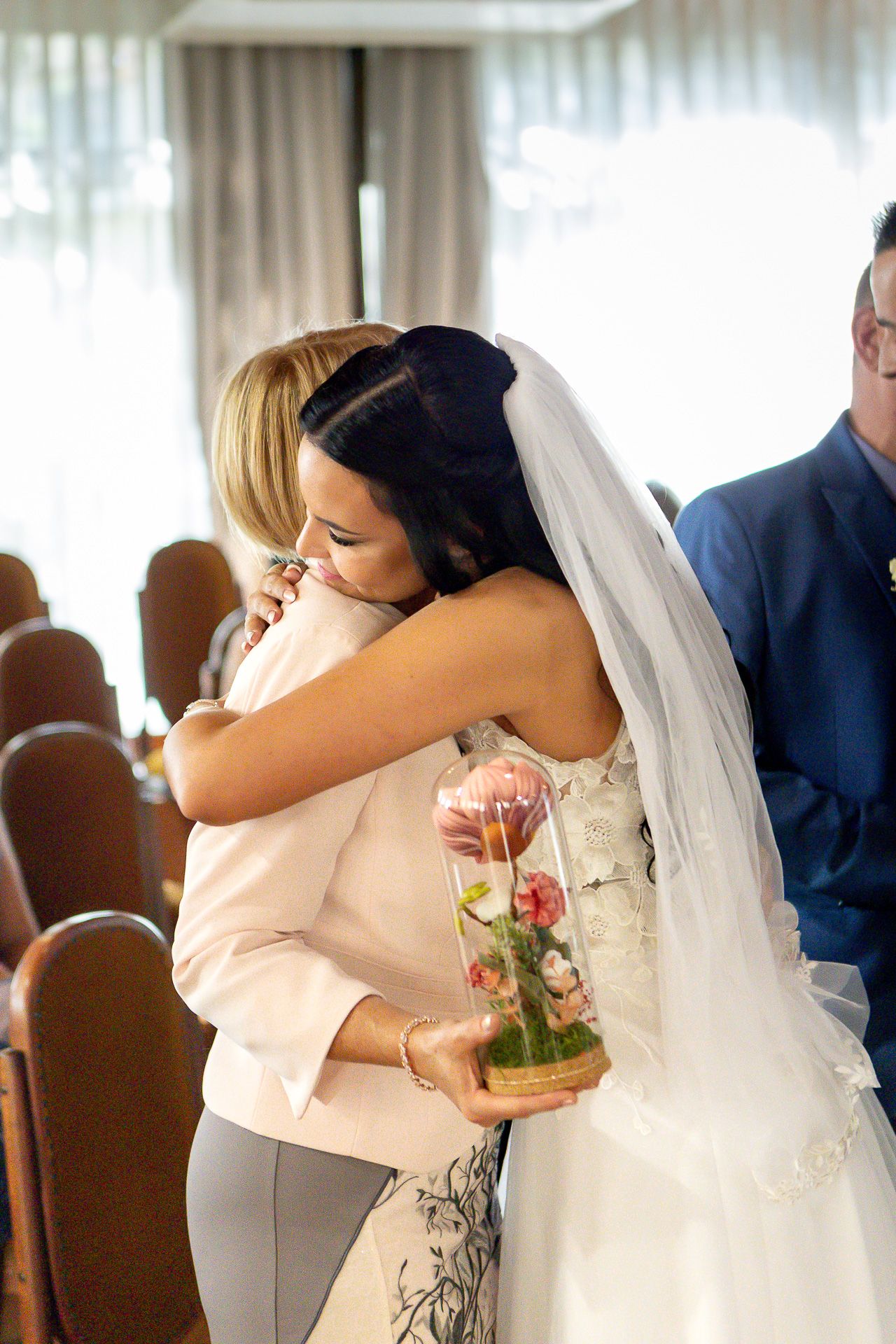A bride in a white wedding dress embraces a guest wearing peach at an indoor wedding reception venue.