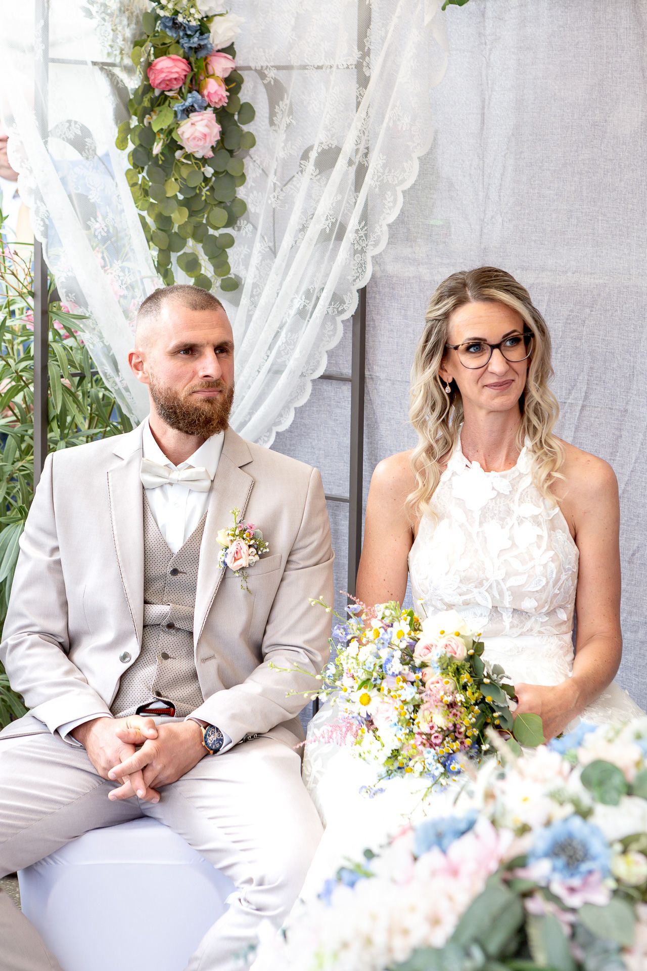 Wedding couple sitting together on white bench with floral decorations and sheer curtains in outdoor setting.