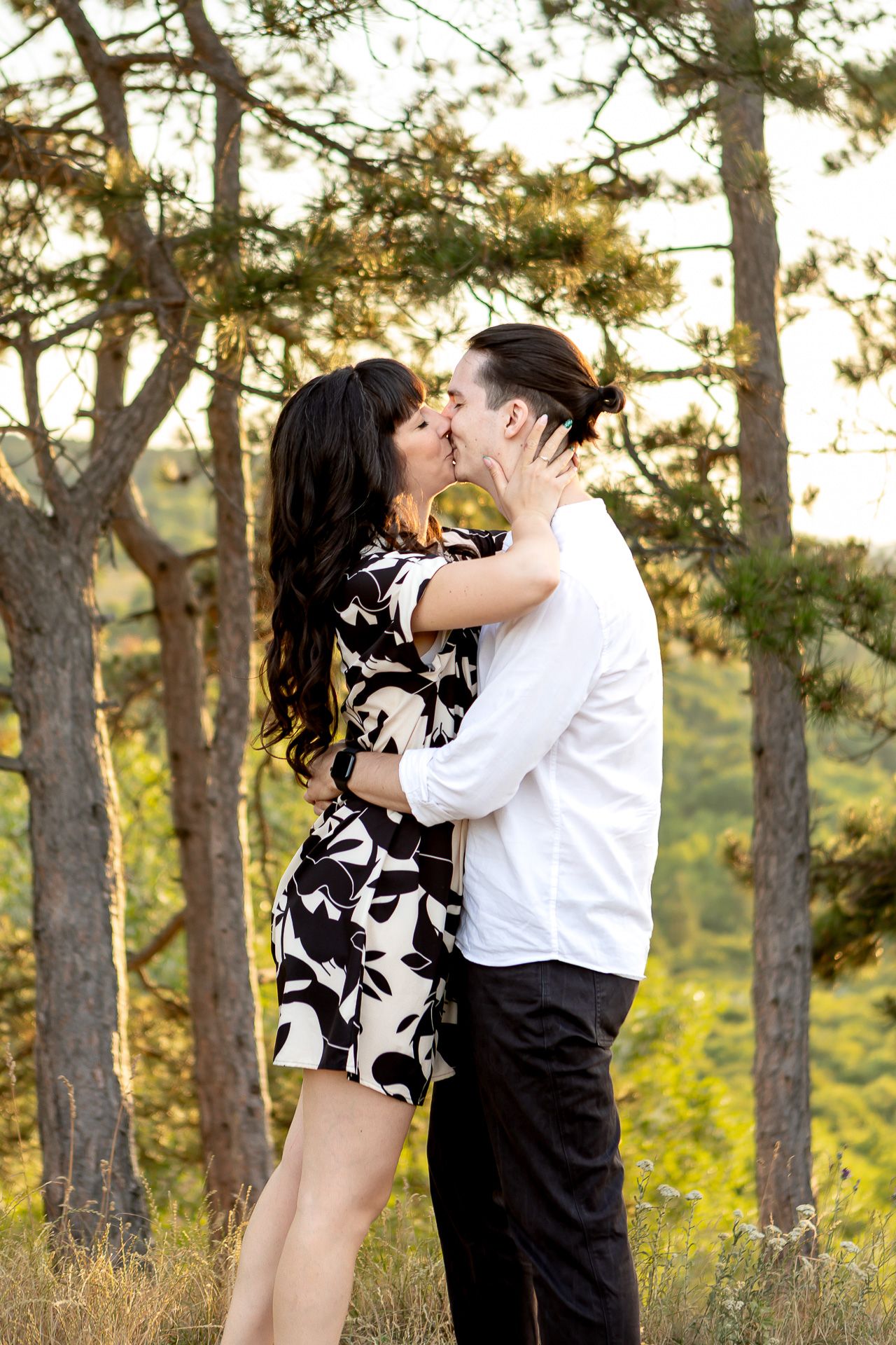 A romantic couple embraces in a scenic forest setting during golden hour with pine trees in the background.