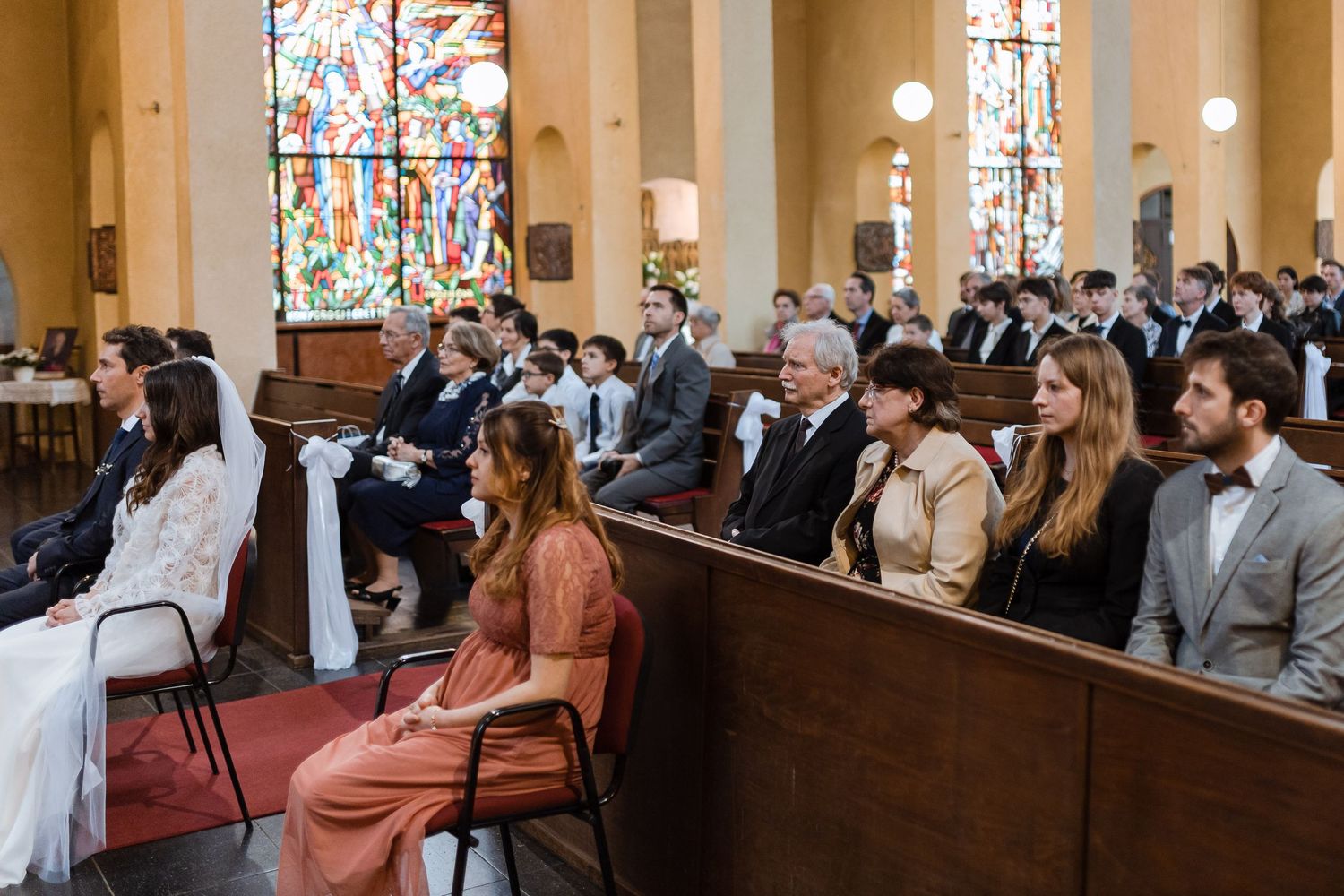 Wedding guests seated in church pews during ceremony with stained glass windows and ornate architecture in background.