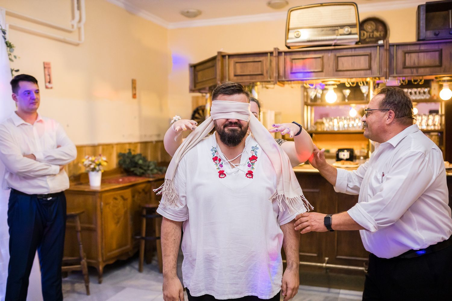 Someone wearing a white embroidered shirt tries on a VR headset while others look on in a room with ambient lighting.