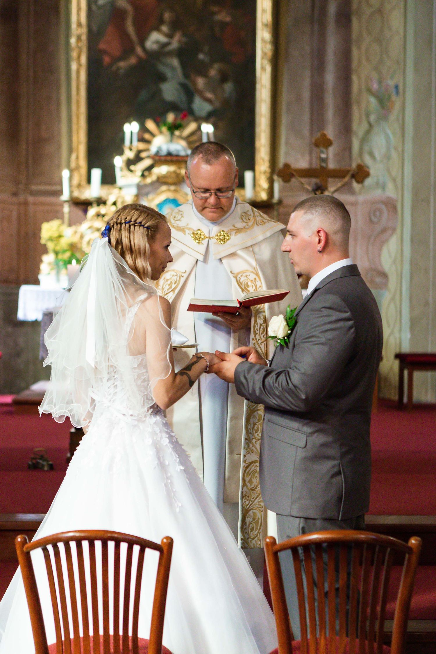 A wedding ceremony takes place inside an ornate church with wooden chairs and golden decor details.