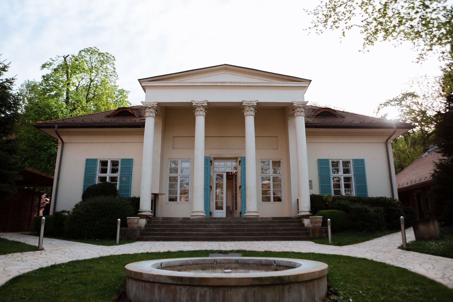 A modern white building with columns and large windows is surrounded by lush green landscaping and trees.